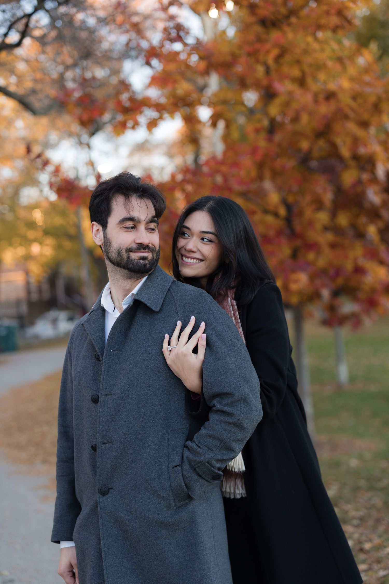 Couple embracing in autumn park, woman smiling in black coat, man in gray coat, colorful fall foliage background.