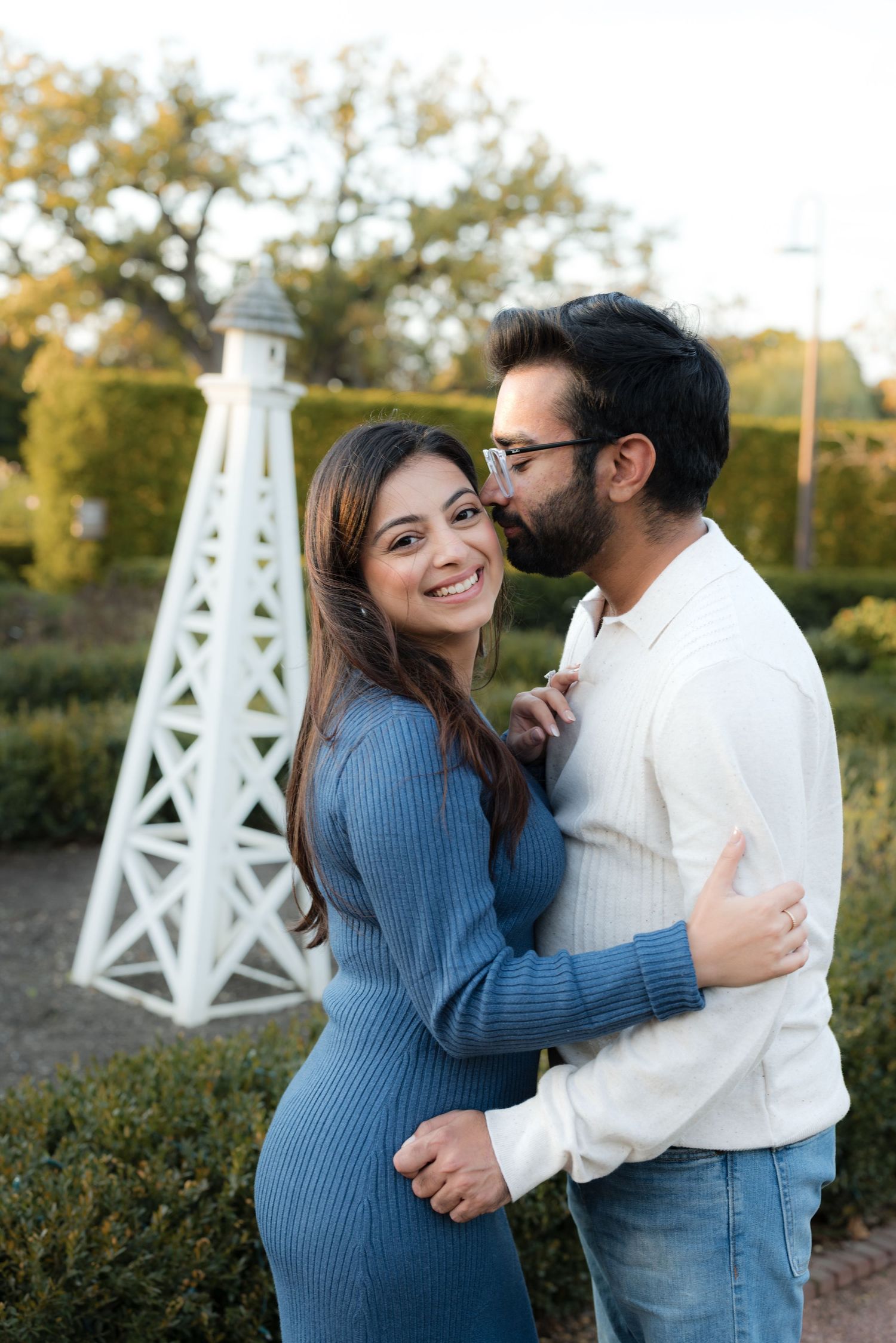 Couple embracing in a garden, woman in blue dress smiling at camera while man kisses her cheek near white lighthouse.