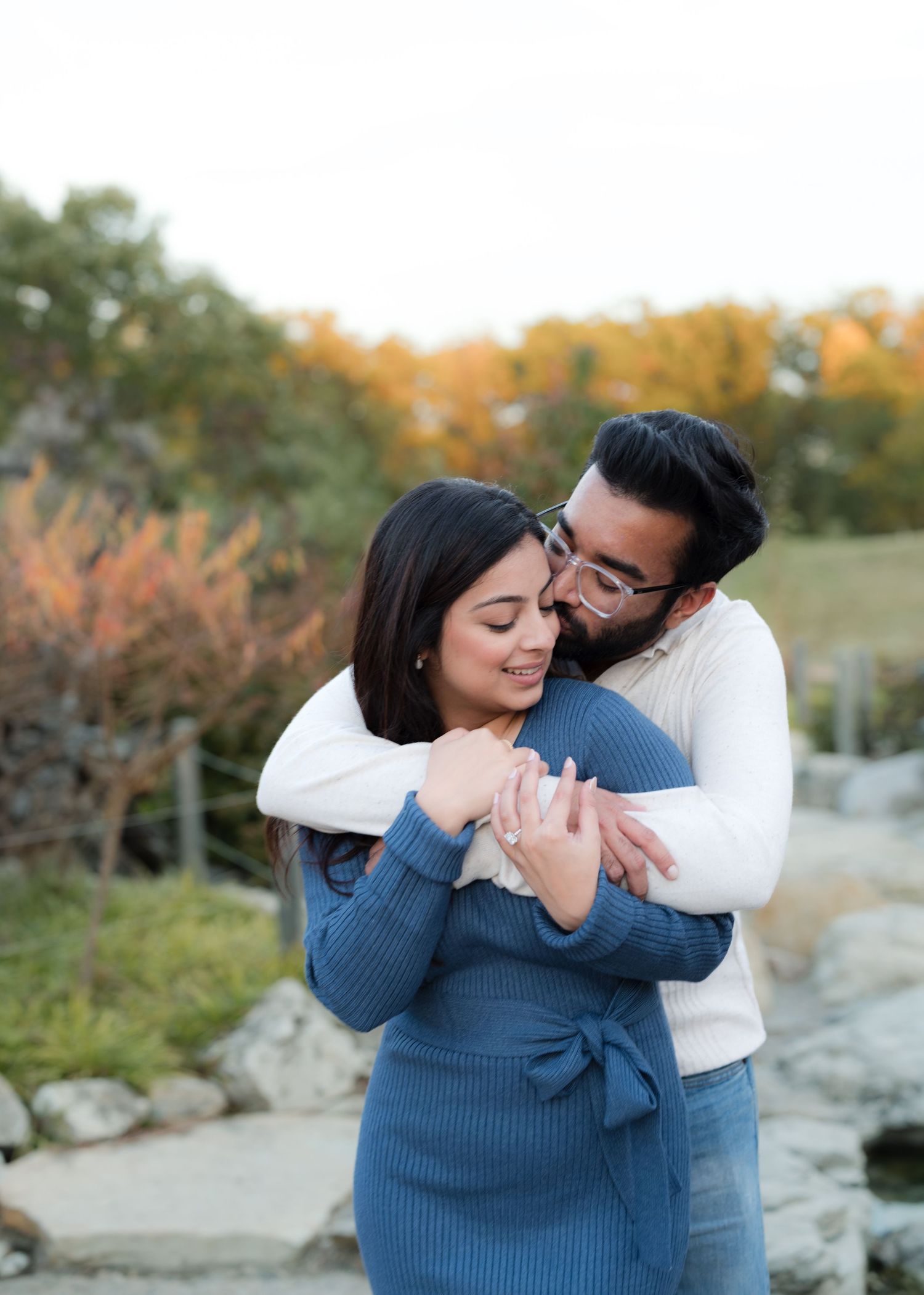 Couple embracing outdoors in autumn, woman in blue dress, man in white sweater, with colorful fall foliage in background.