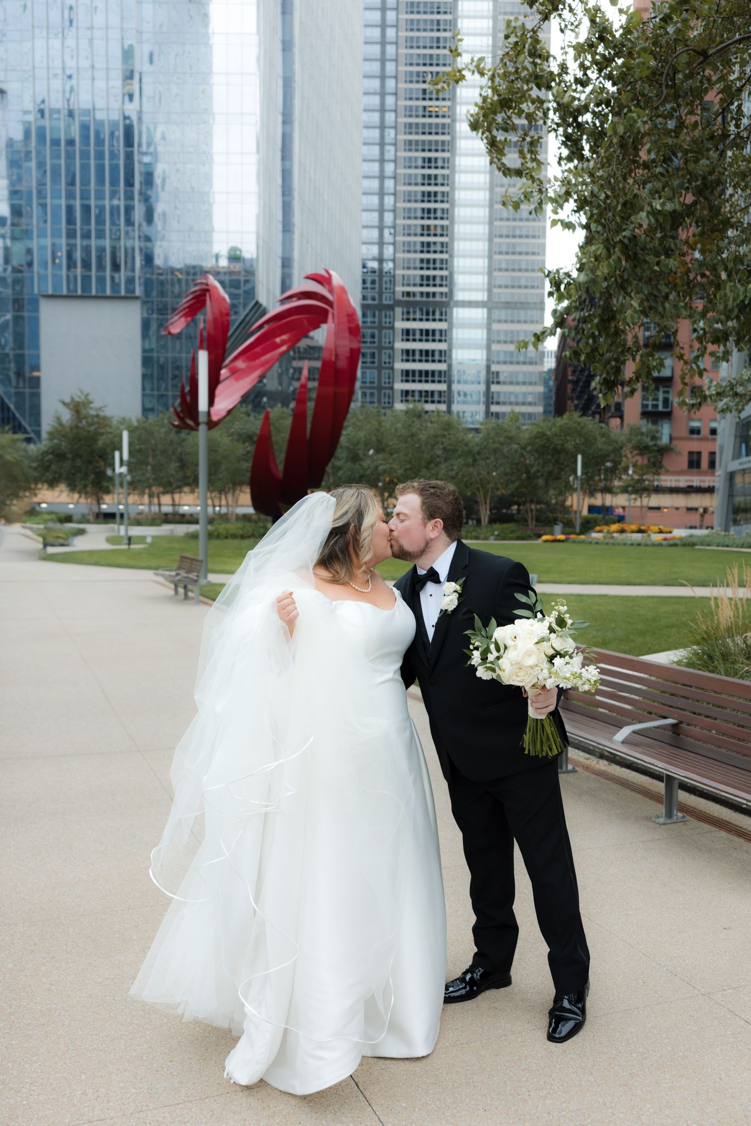 Wedding couple poses playfully against urban backdrop with red wind-blown veil.