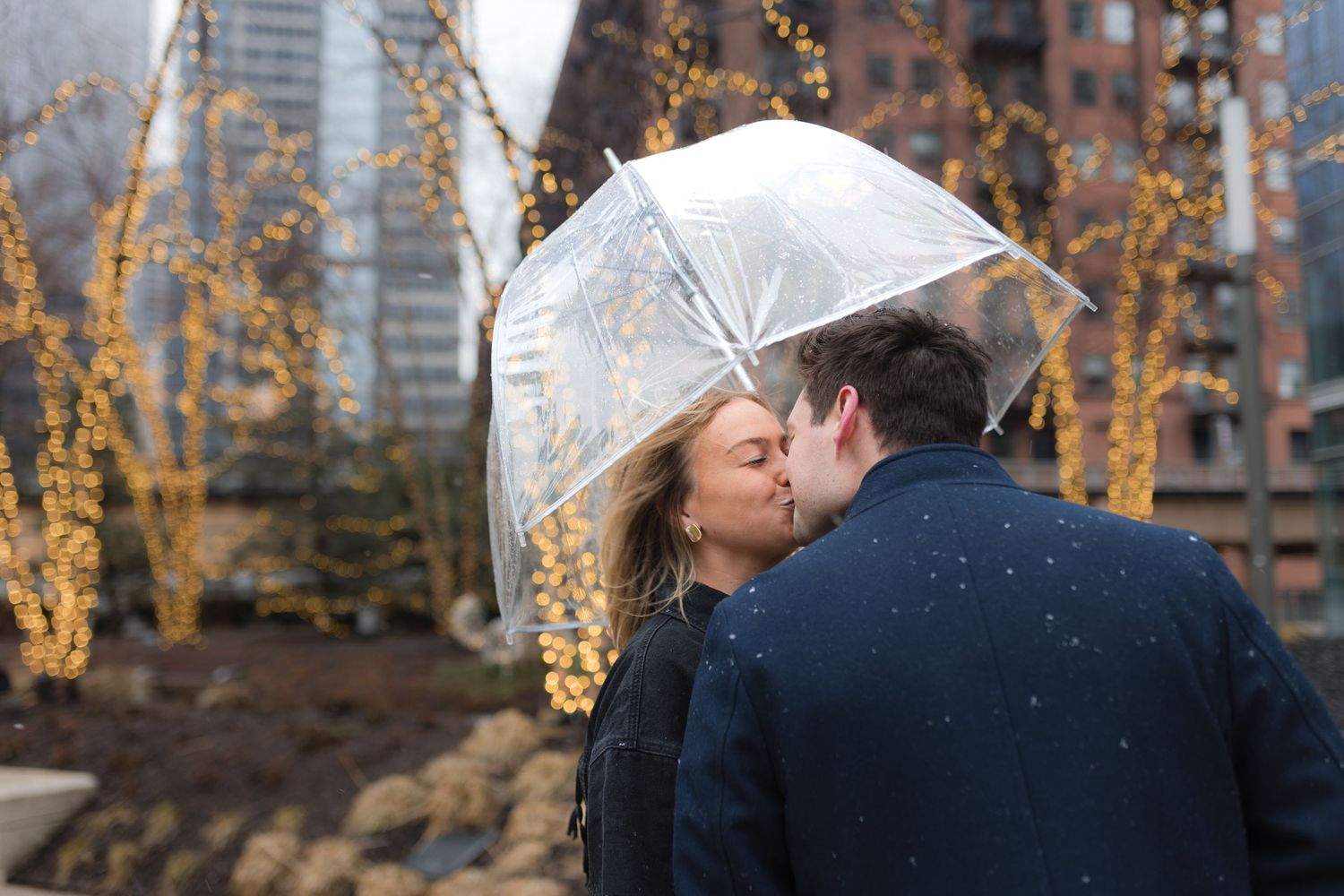 Couple sharing a romantic kiss under a clear umbrella in the rain with city lights and buildings in the background.