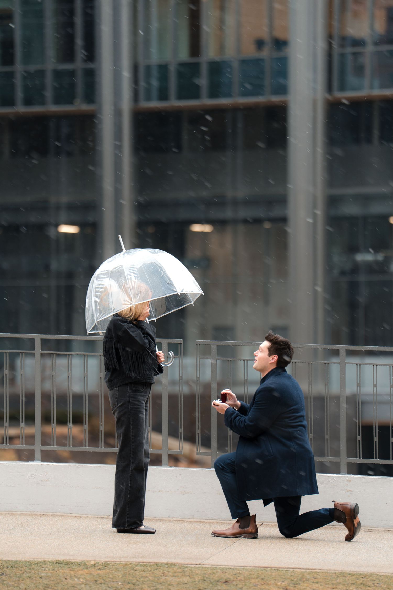Man proposing on one knee to woman holding clear umbrella in snow outside modern building.