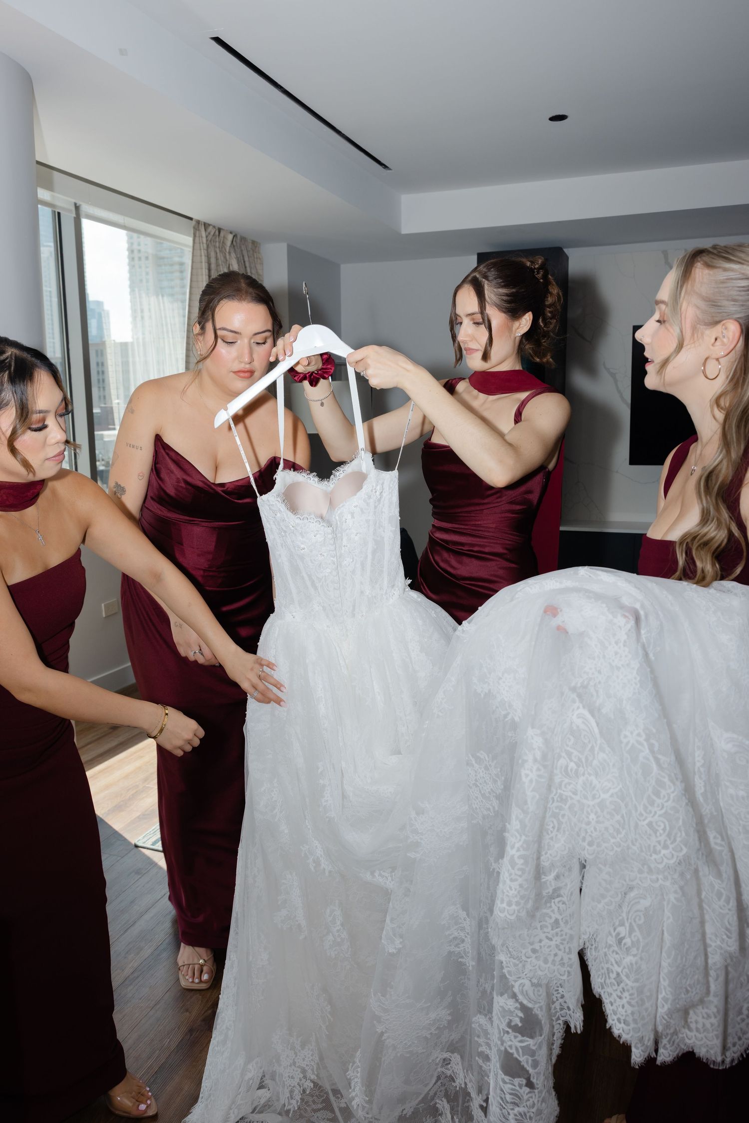 Bridesmaids in burgundy dresses help prepare a white lace wedding gown in a modern hotel room.