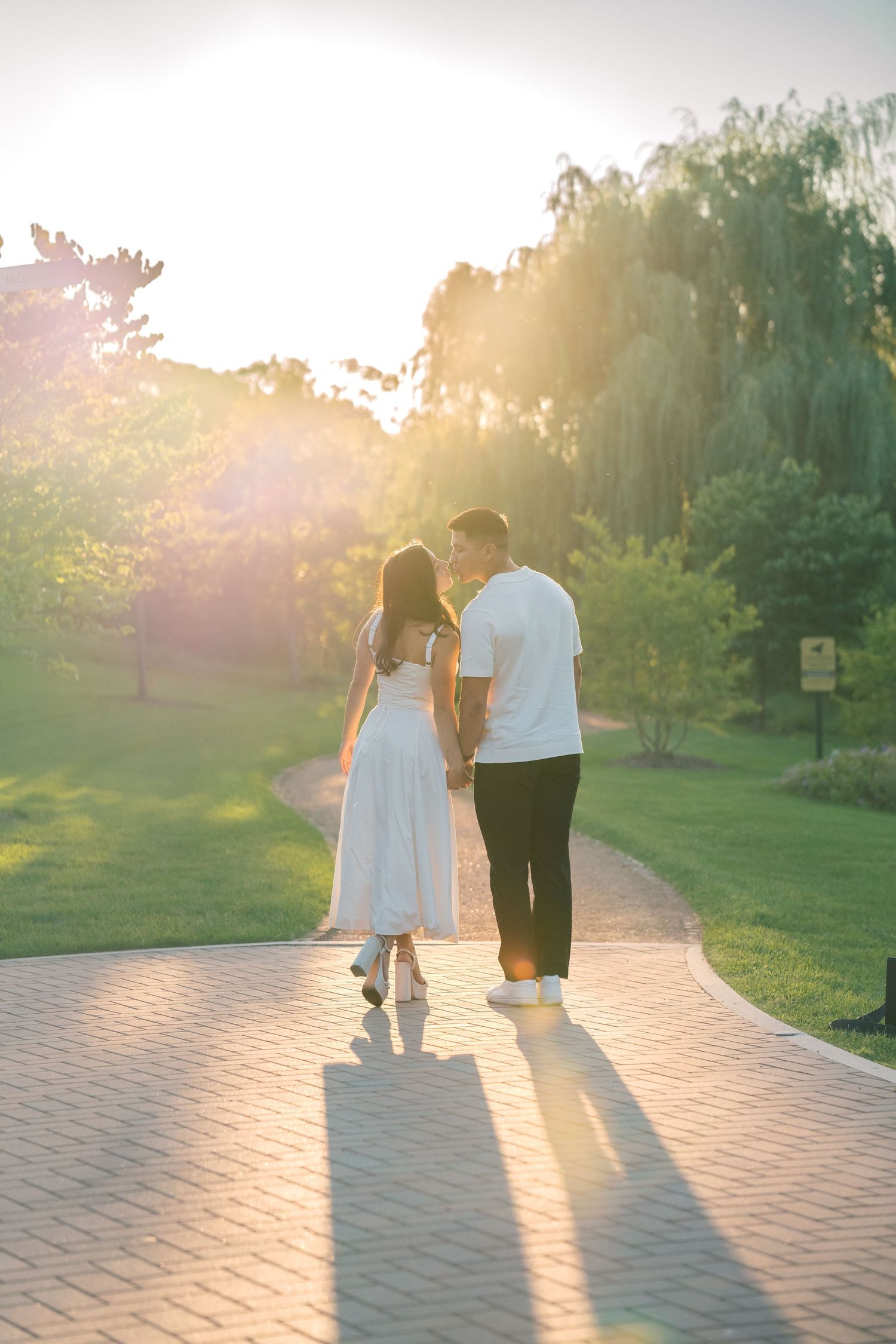 Couple holding hands walking on a brick path at golden hour sunset in a lush green park.