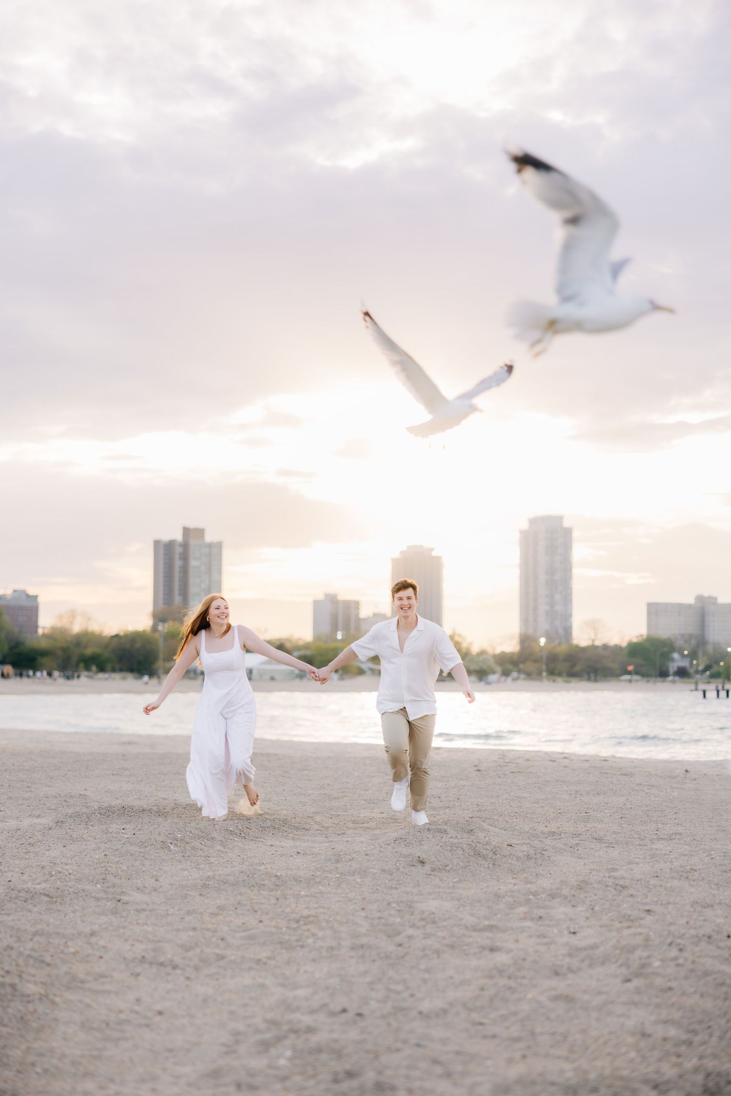 Couple in white clothing dancing on beach with seagulls flying overhead at sunset.
