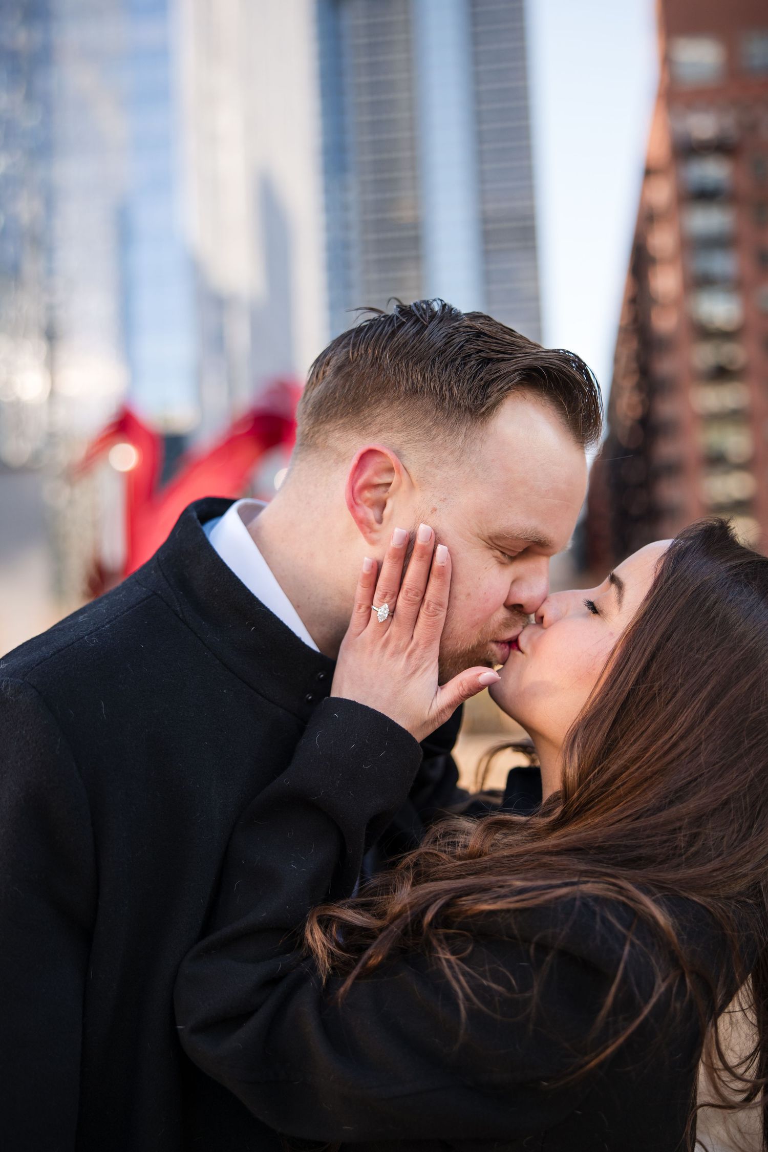 Couple sharing a romantic kiss outdoors in an urban setting, woman wearing engagement ring, city buildings in background.