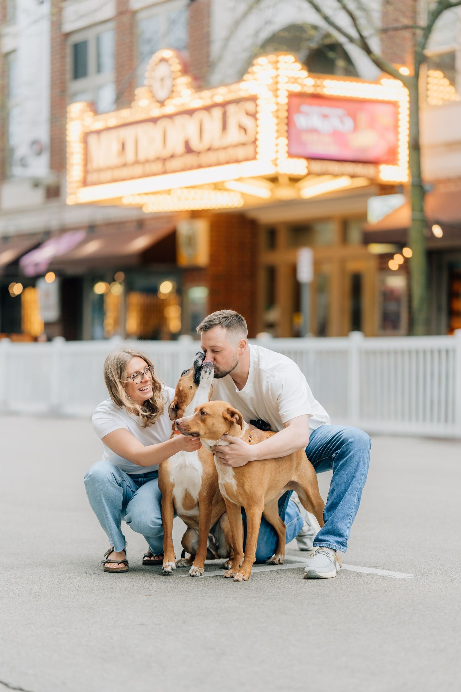 Couple in white tees and jeans poses with two brown dogs in front of Metropolis Theatre marquee.