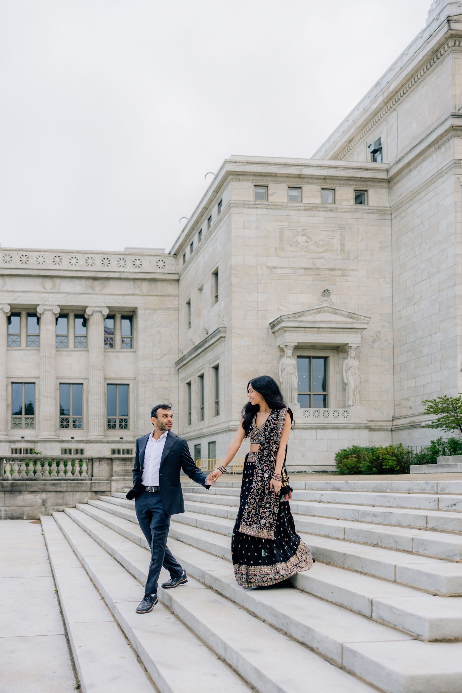 Couple in formal attire holding hands on stone steps outside a grand neoclassical building.