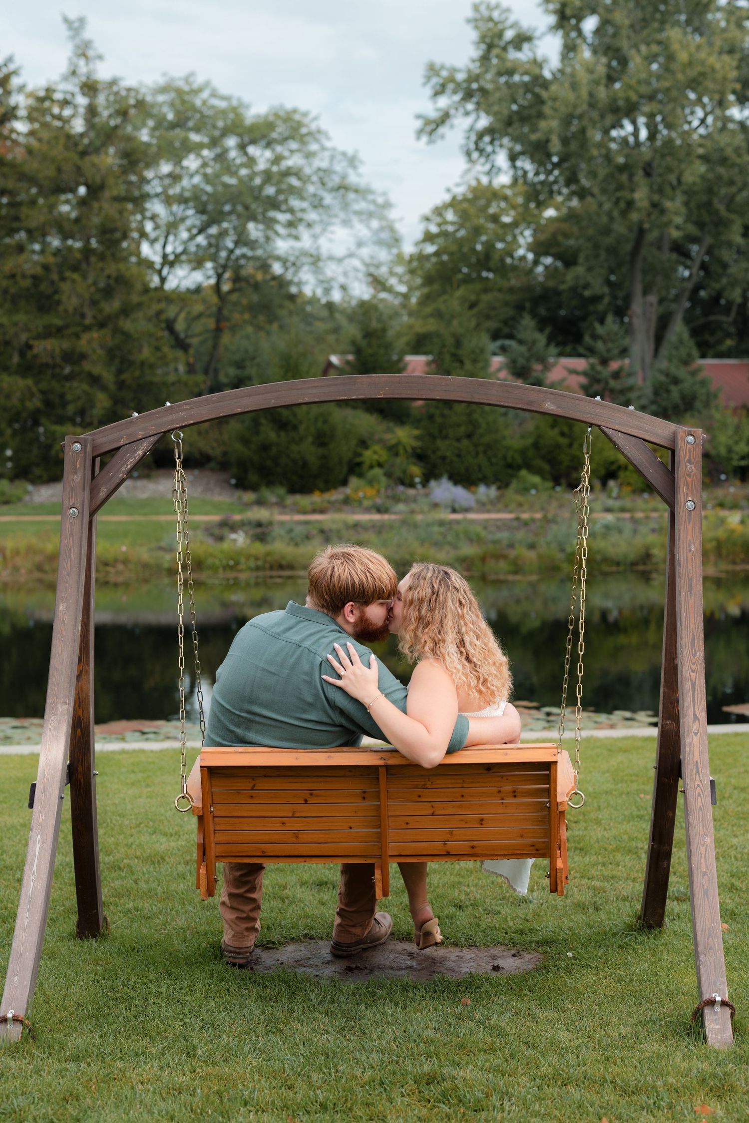 Couple sharing a kiss on a wooden garden swing bench beside a serene pond surrounded by lush green trees.