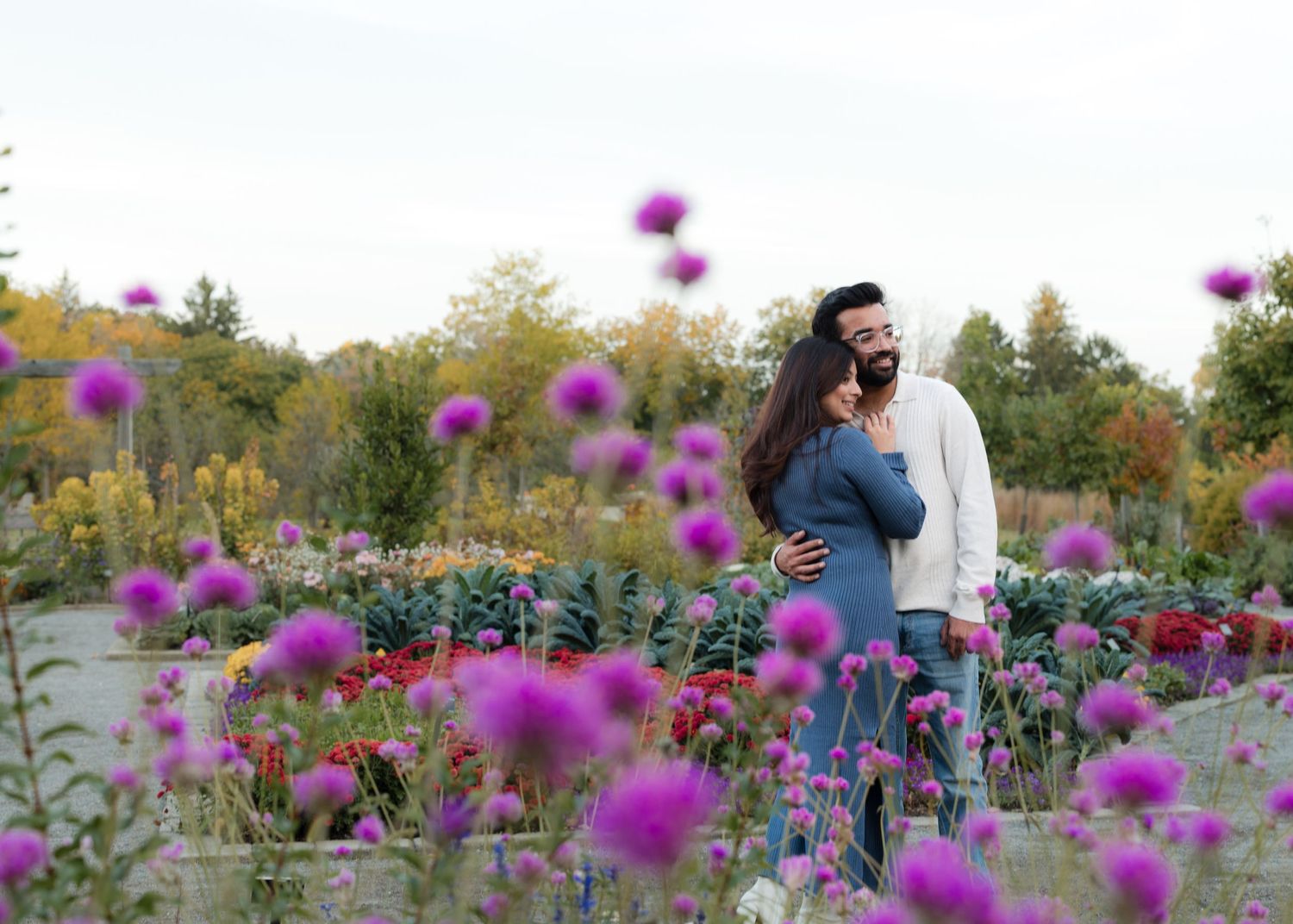 Couple embracing in a vibrant flower garden with purple blooms in the foreground, wearing masks outdoors.
