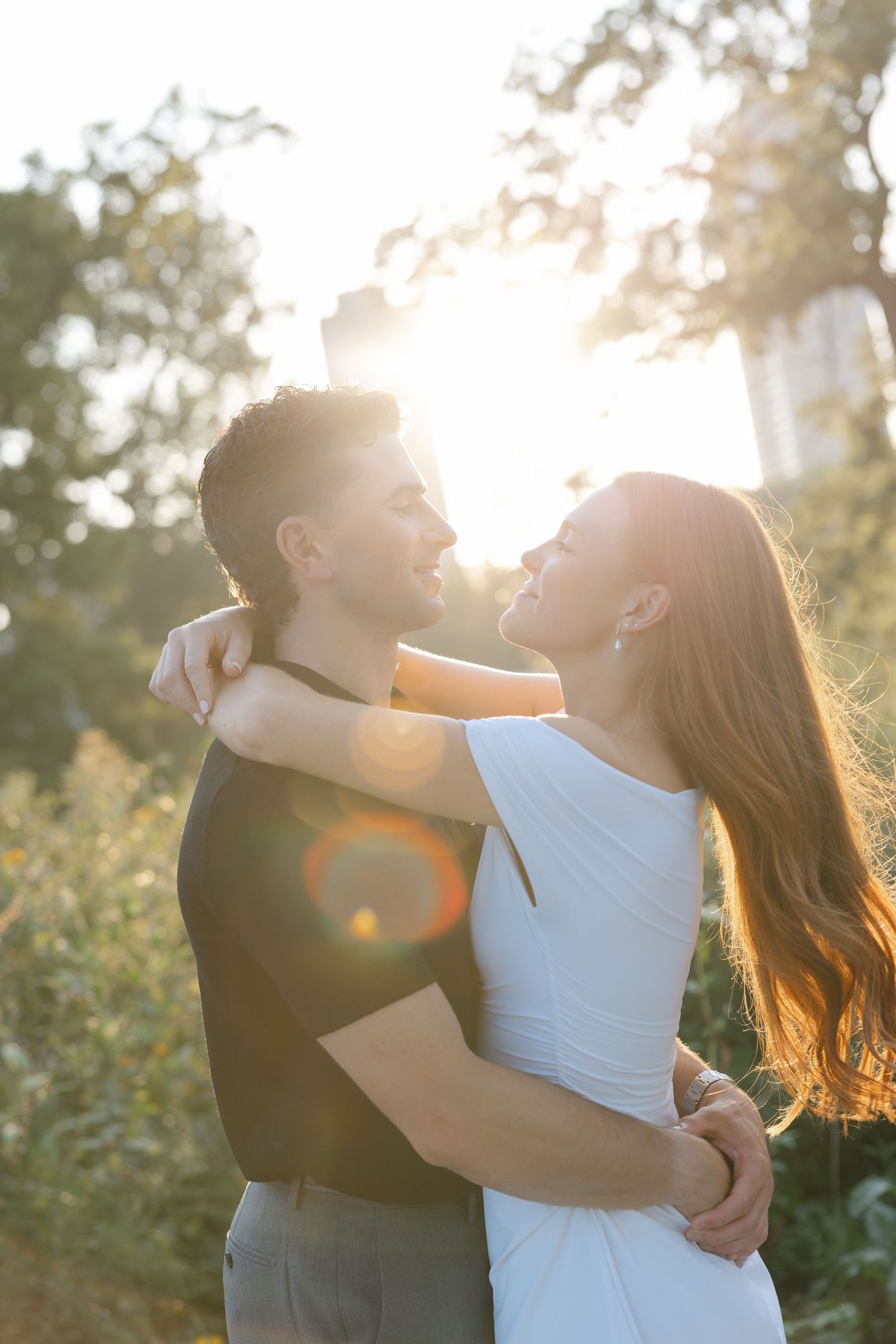 Couple embracing in golden sunlight, woman in white dress with long red hair, surrounded by lush green trees.