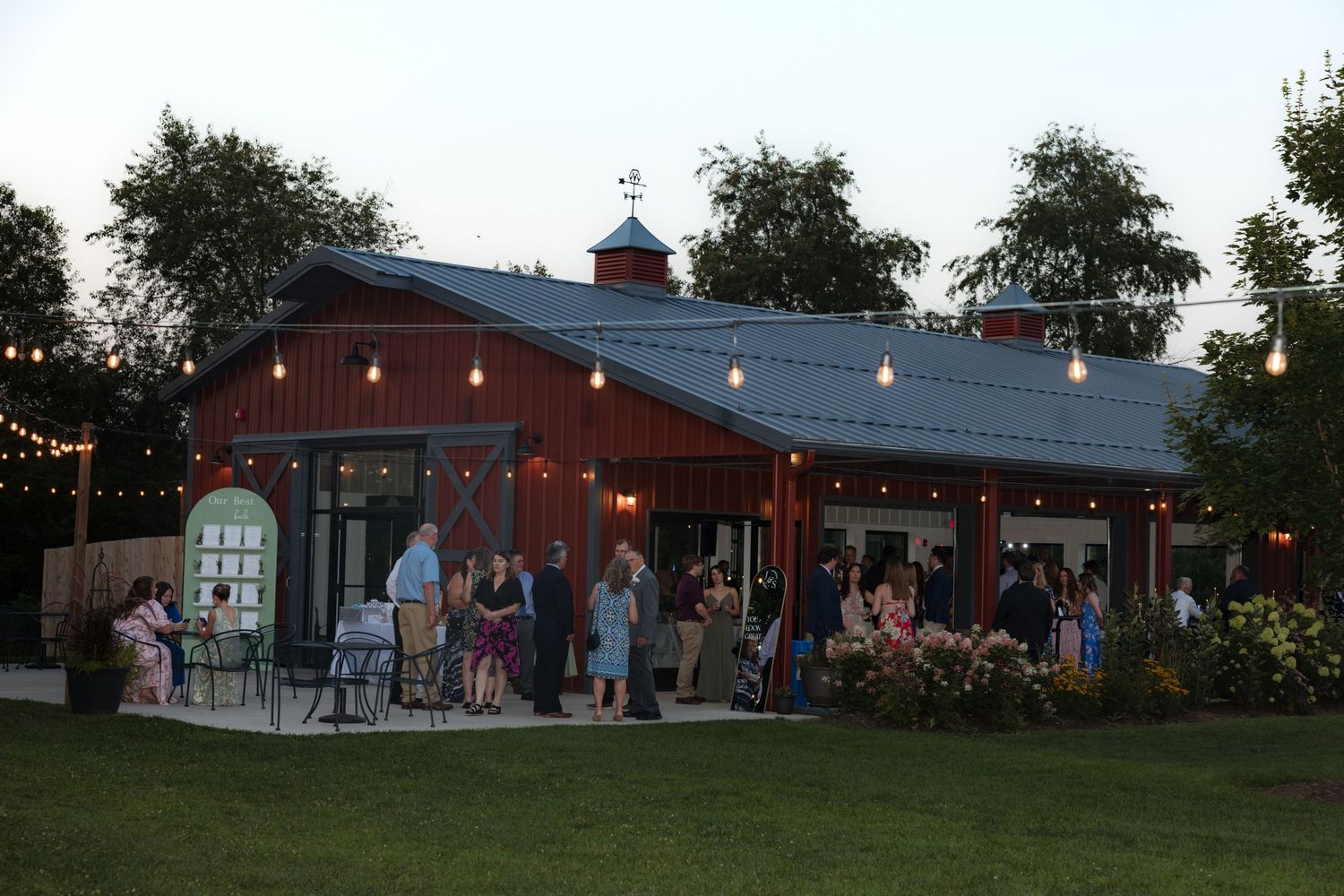 Panoramic view of outdoor wedding reception venue with red barn and string lights at dusk.