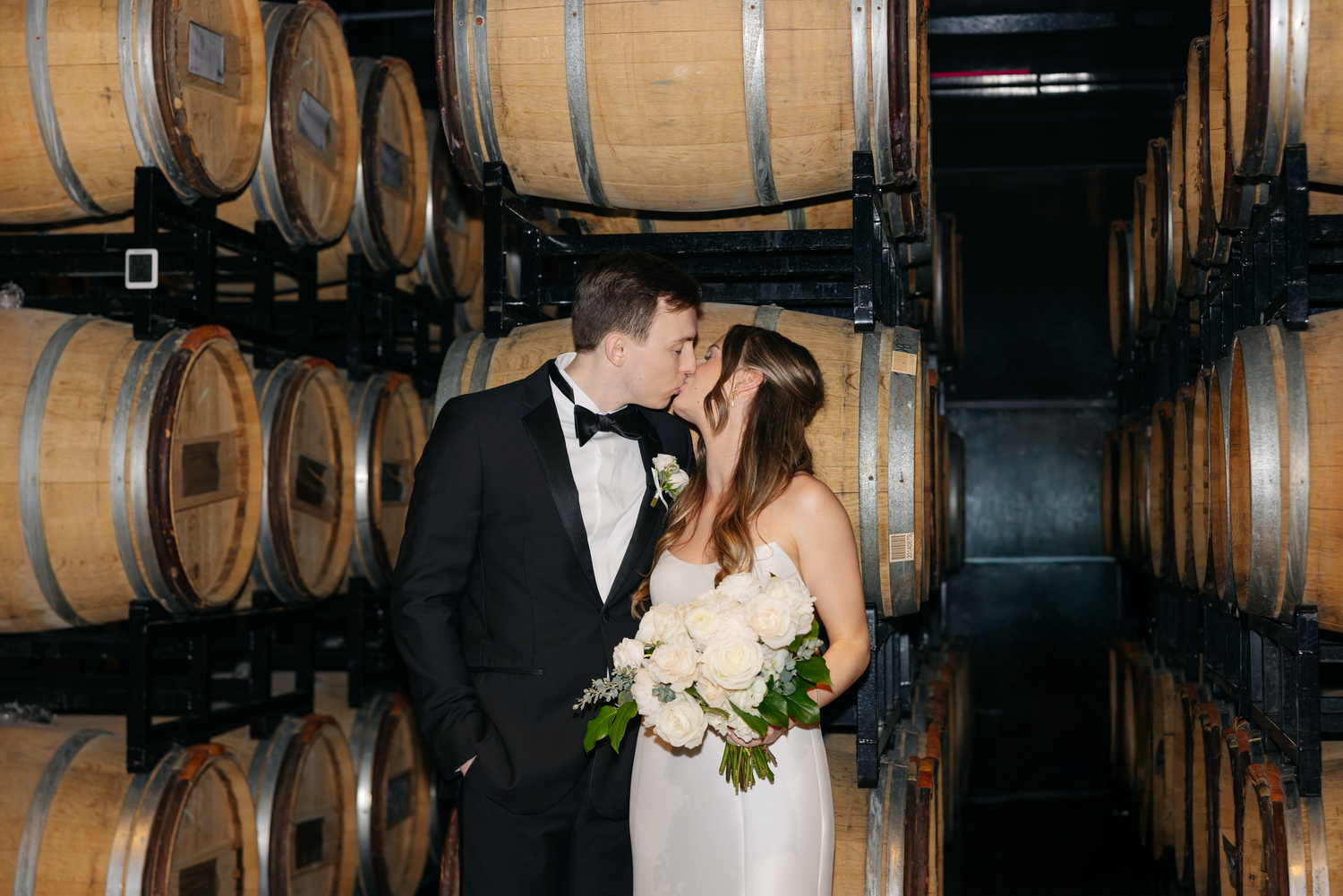 Bride and groom share a romantic kiss surrounded by oak wine barrels in a winery cellar wedding portrait.