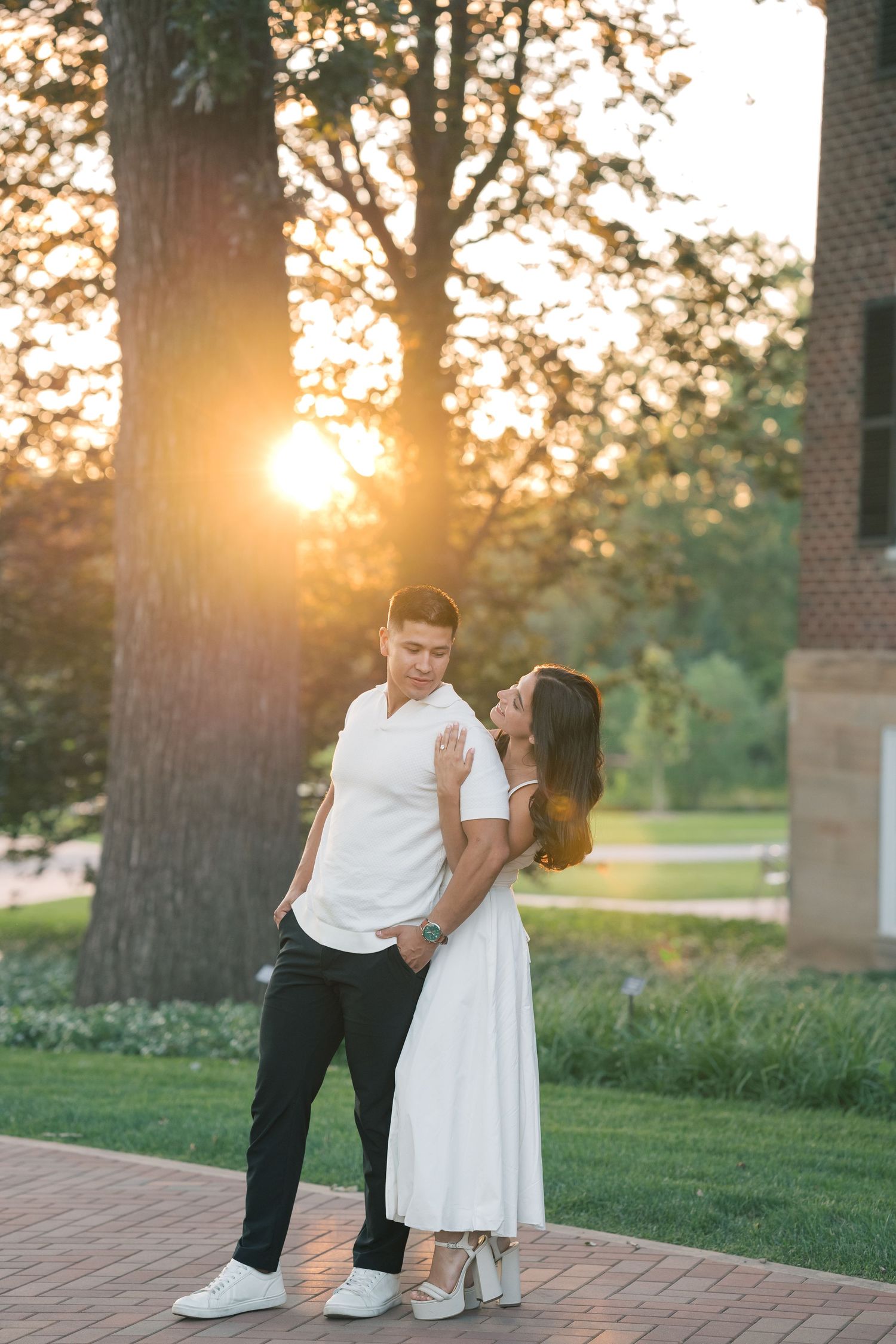 Couple embracing at golden hour sunset in a park, woman in white dress, man in white polo and black pants.