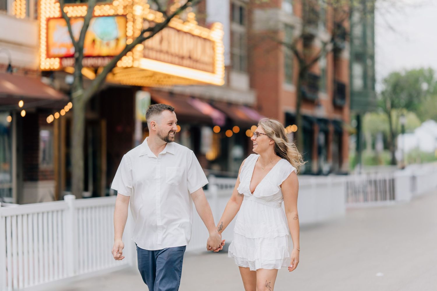 Couple in white outfits holding hands and smiling while walking along a charming downtown street at dusk.
