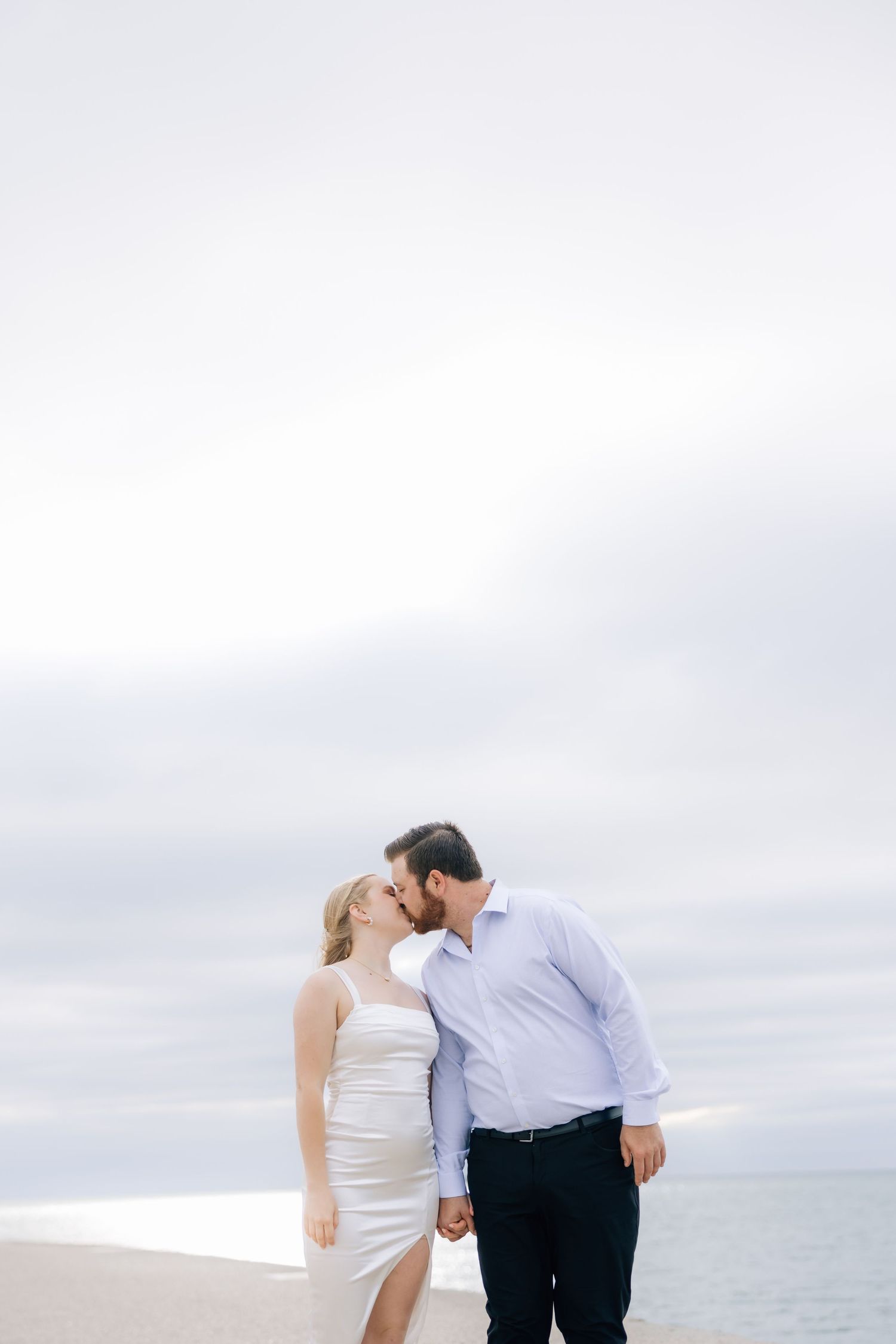 A couple shares a romantic kiss on a beach, the woman in a white dress holding hands with a man in a light blue shirt.