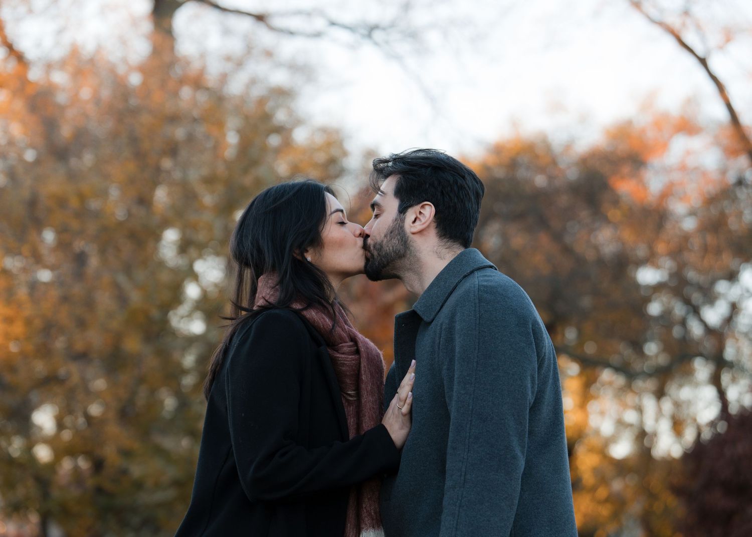 Couple sharing a romantic kiss outdoors in autumn park with colorful fall foliage in the background.