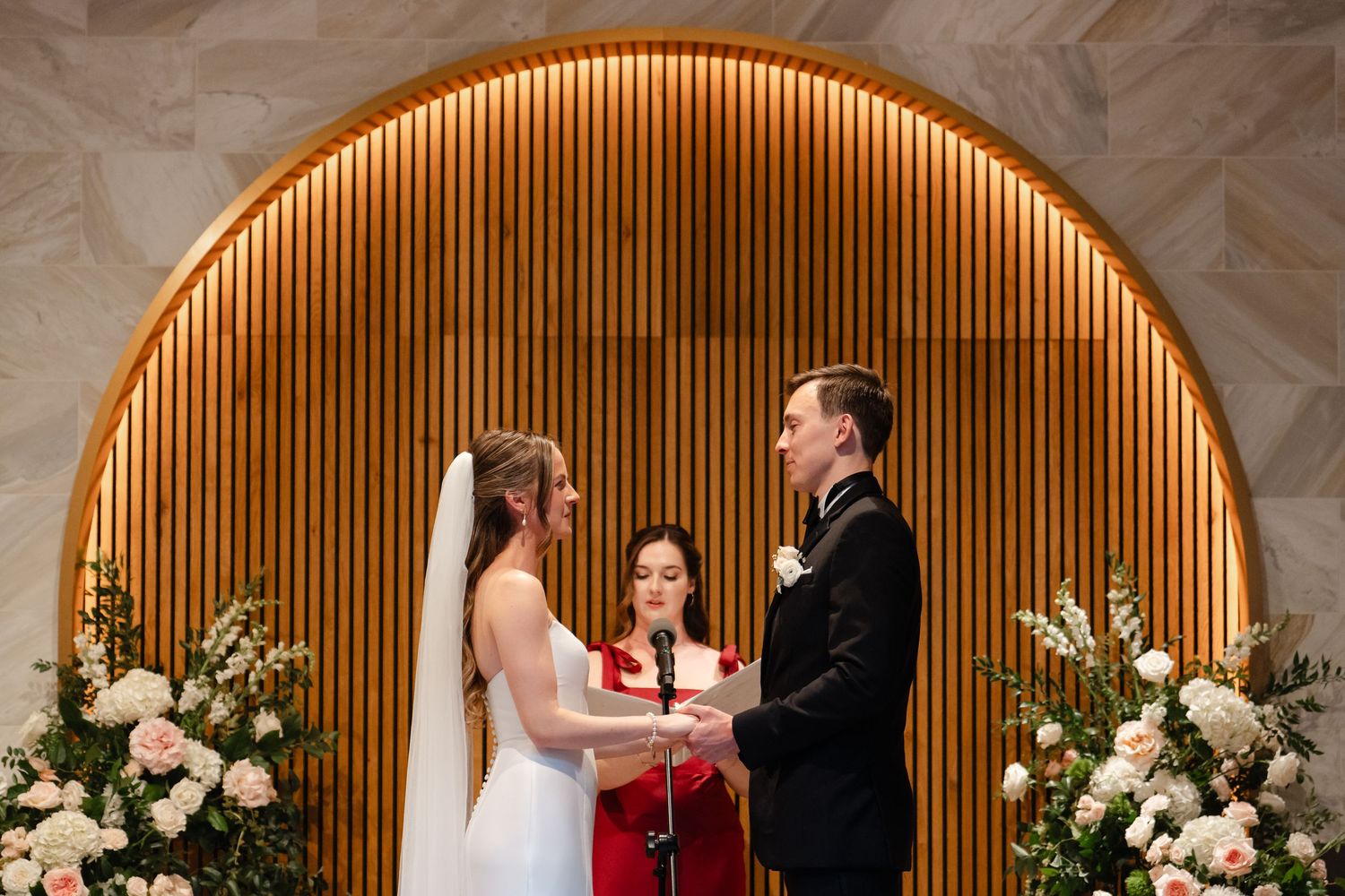 Bride and groom exchange vows at elegant wedding ceremony with arched wood backdrop and floral arrangements.