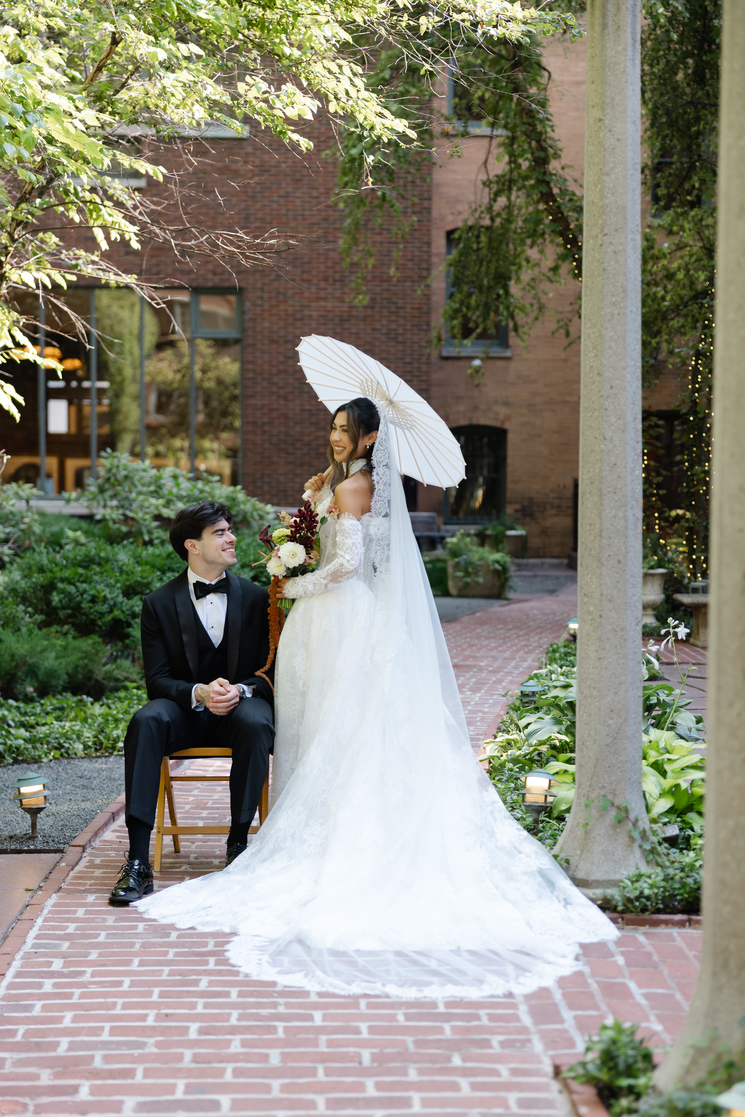 Bride holding white parasol stands beside groom seated in chair on brick garden path during outdoor wedding portrait.