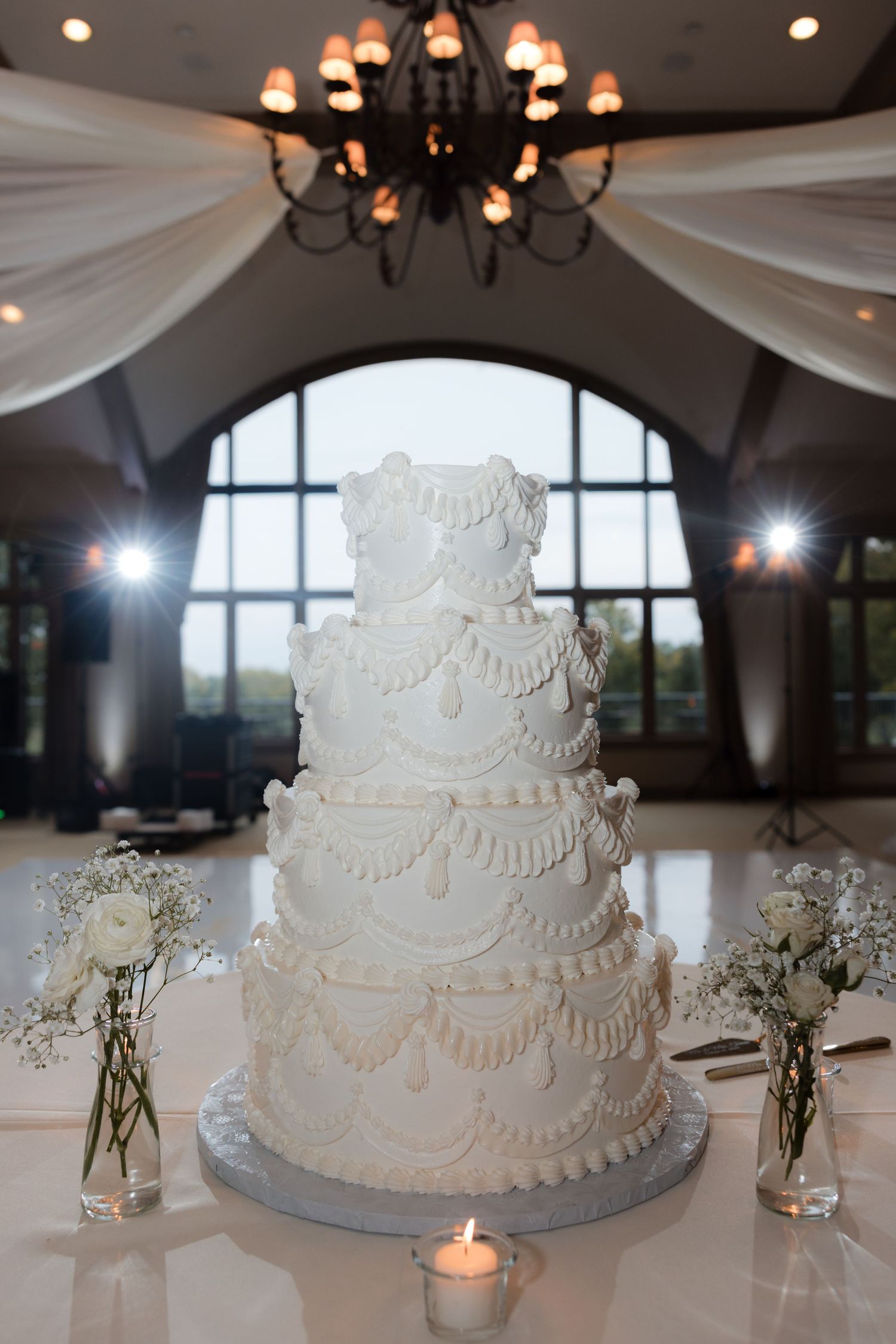 Elegant white tiered wedding cake with intricate piping displayed under chandelier in reception venue.
