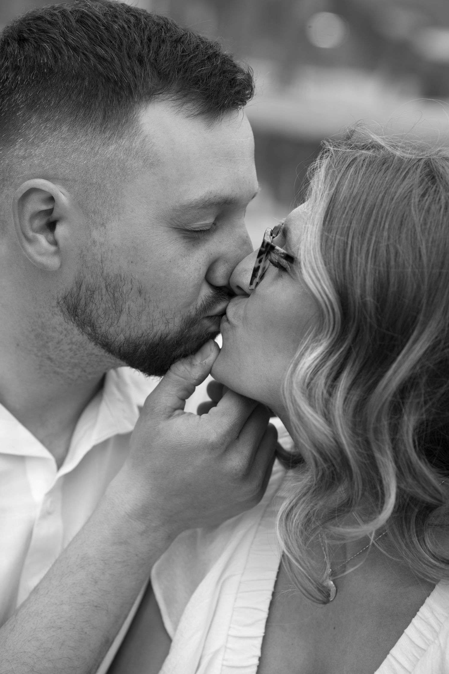 Black and white photo of a couple sharing a romantic kiss, woman wearing glasses with wavy hair.
