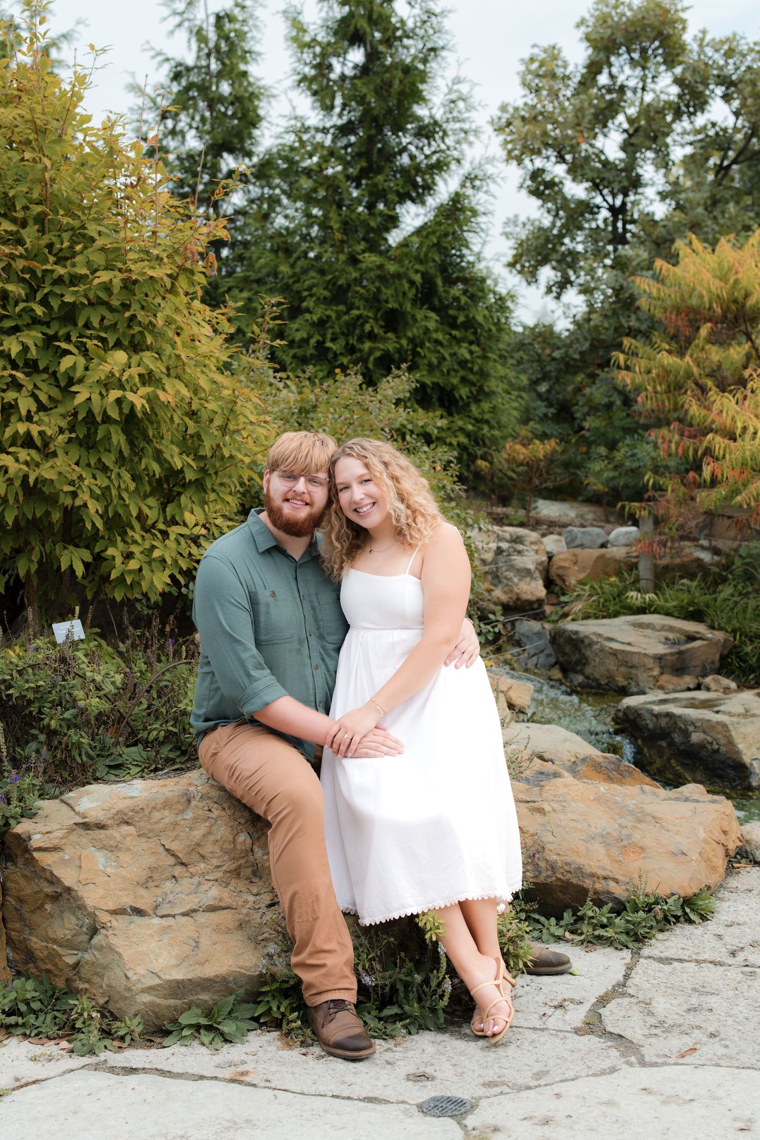 A couple sits on rocks in a garden, woman in white dress leaning on man in green shirt, surrounded by lush greenery.