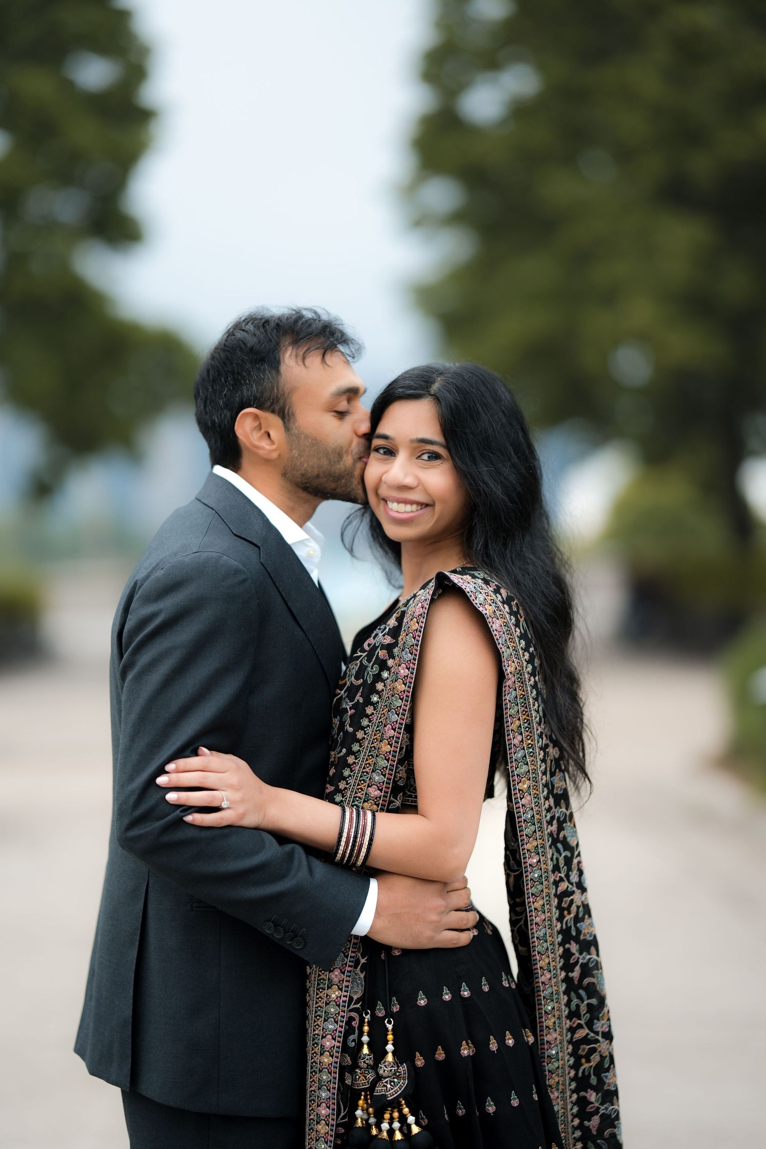 Indian couple in traditional and formal attire embracing outdoors, man kissing woman's cheek as she smiles.