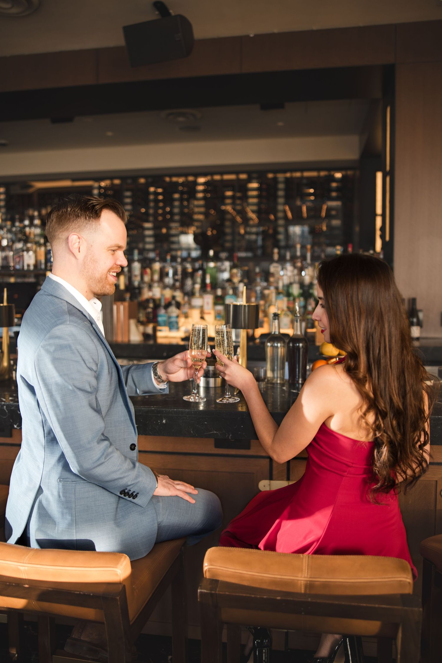 Couple toasting champagne glasses at an upscale bar, man in gray suit and woman in red dress smiling.