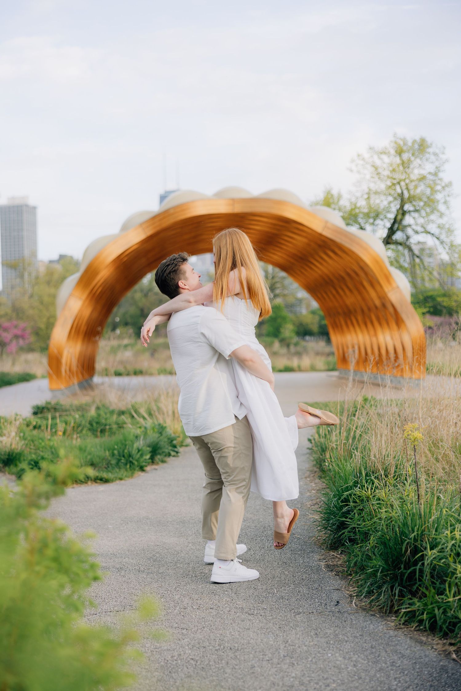 Couple embracing on a park path with a golden arch sculpture and city skyline in the background.