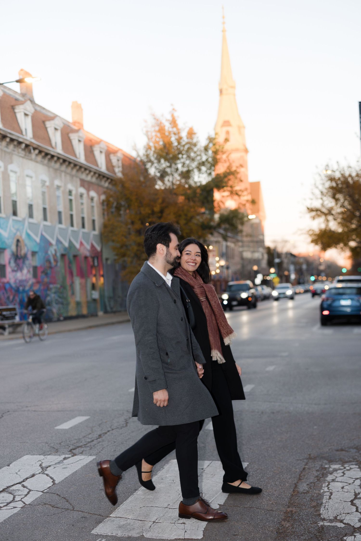 Couple walking hand in hand across a crosswalk in an urban setting with a church steeple and colorful mural in the background.