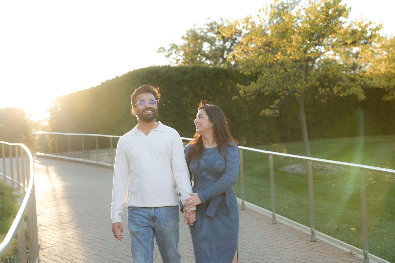 Couple holding hands walking on a sunlit bridge path surrounded by lush green trees at golden hour.
