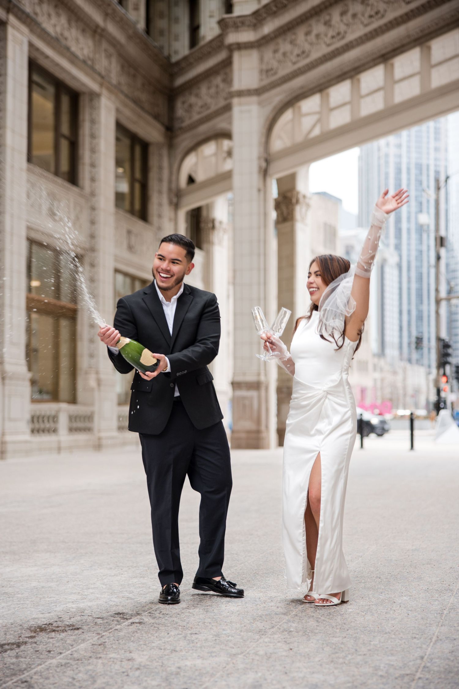 Joyful couple in wedding attire celebrating outdoors, popping champagne bottle on urban city street.