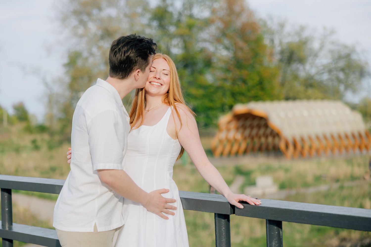 Couple embracing on a bridge outdoors, woman in white dress smiling, man in white shirt, green landscape background.