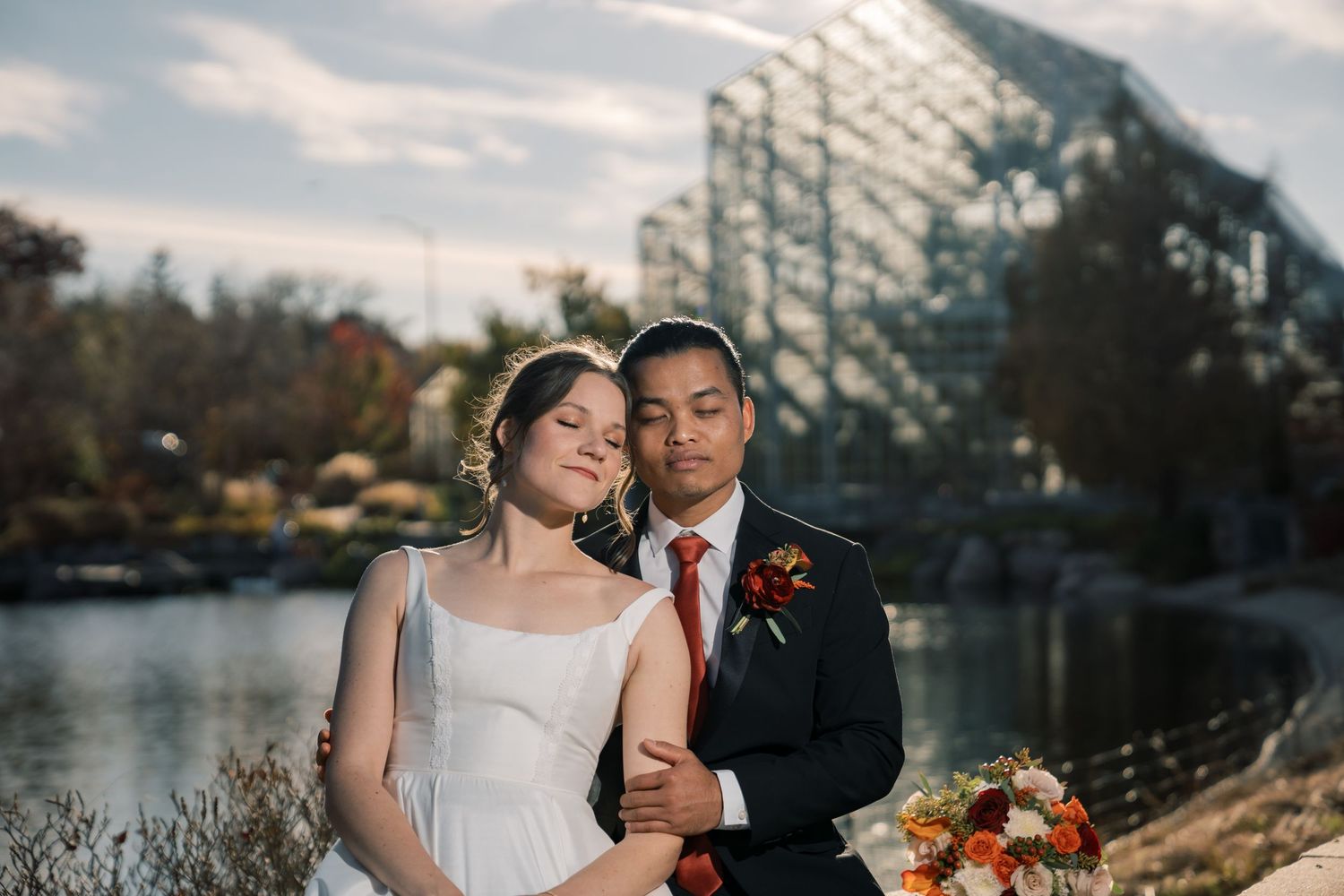 Wedding couple embracing by waterfront with city skyline in background.