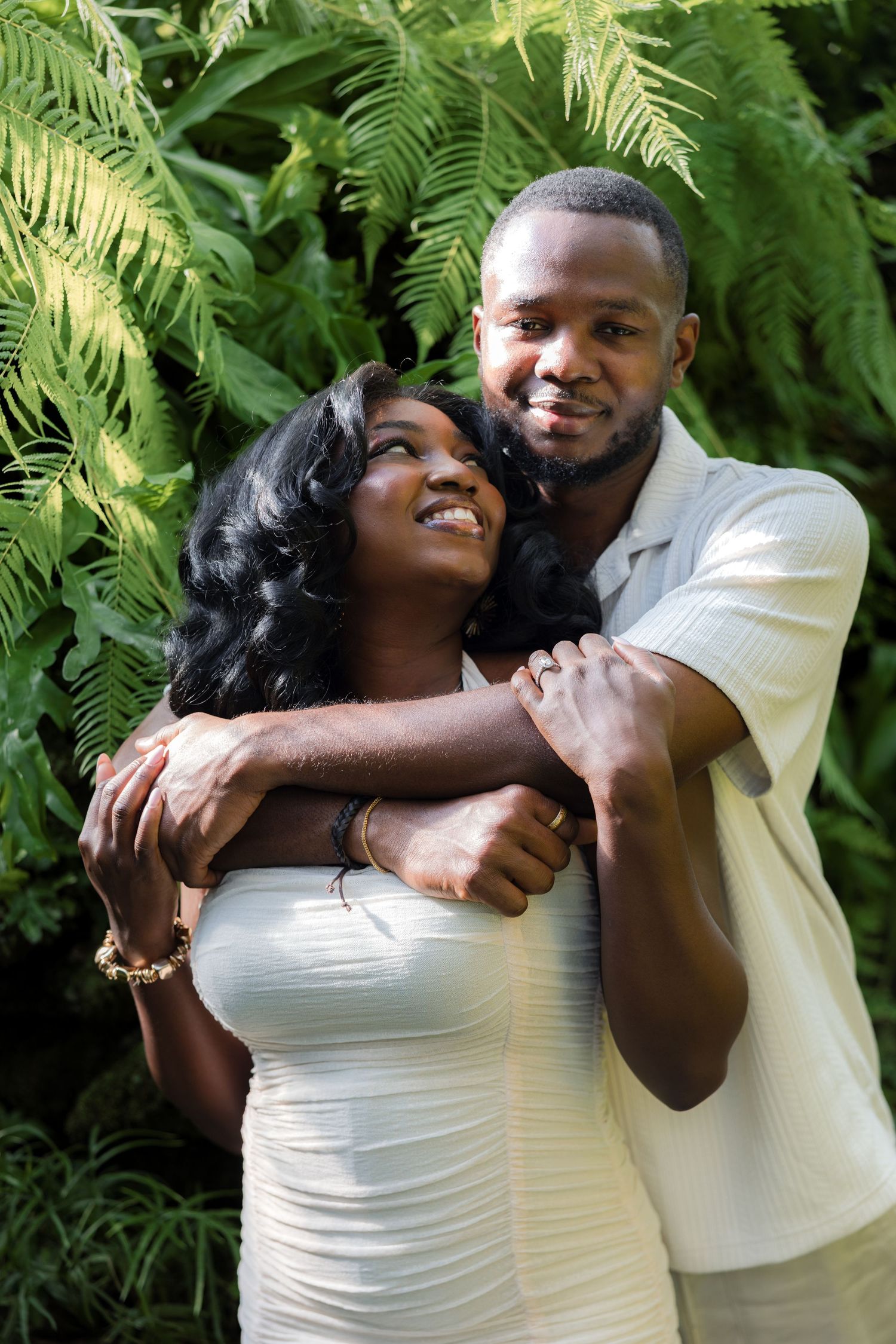 A joyful Black couple embraces outdoors, wearing white outfits, surrounded by lush green tropical ferns.