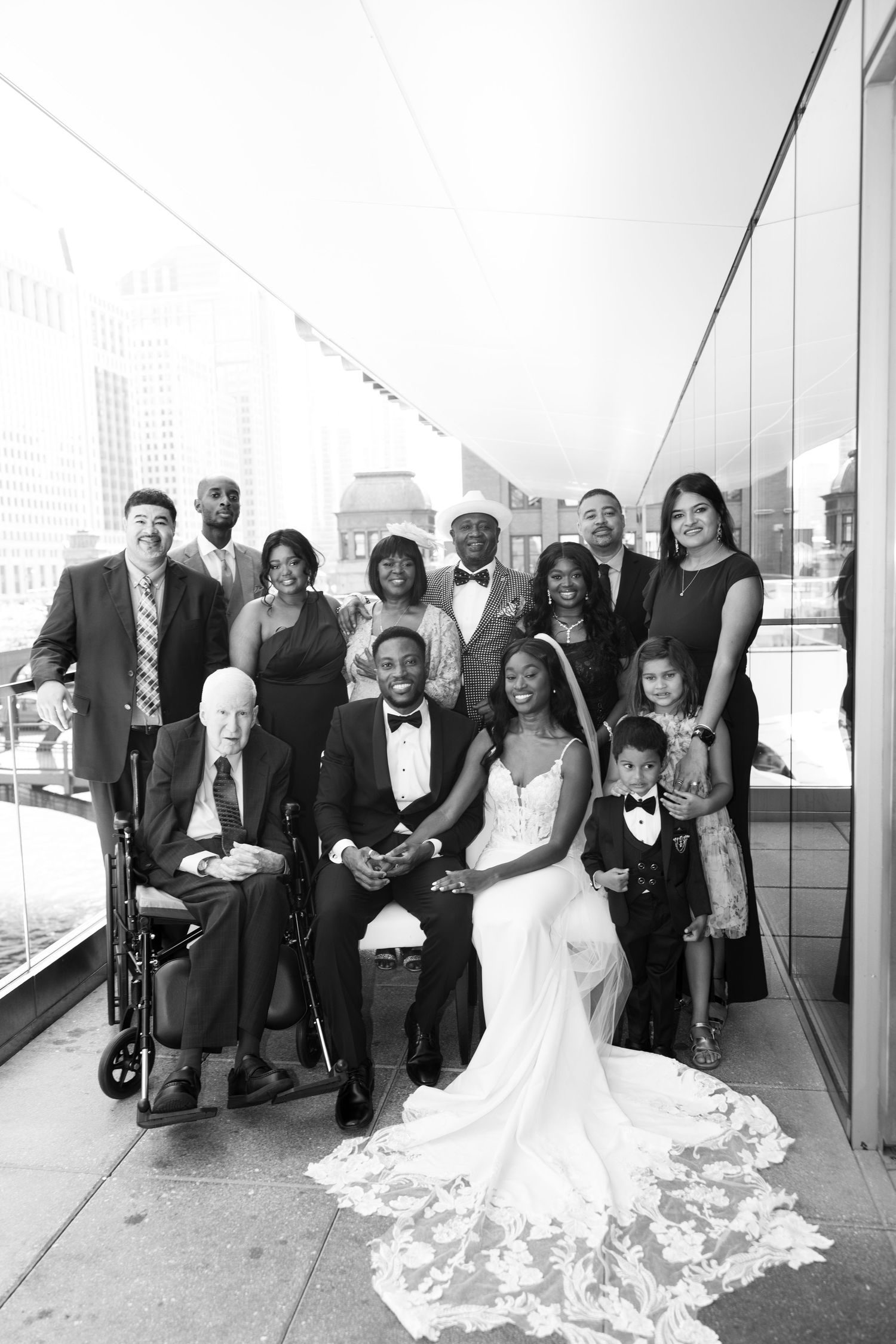 Wedding party in formal attire gathers for group photos on modern balcony with city views.