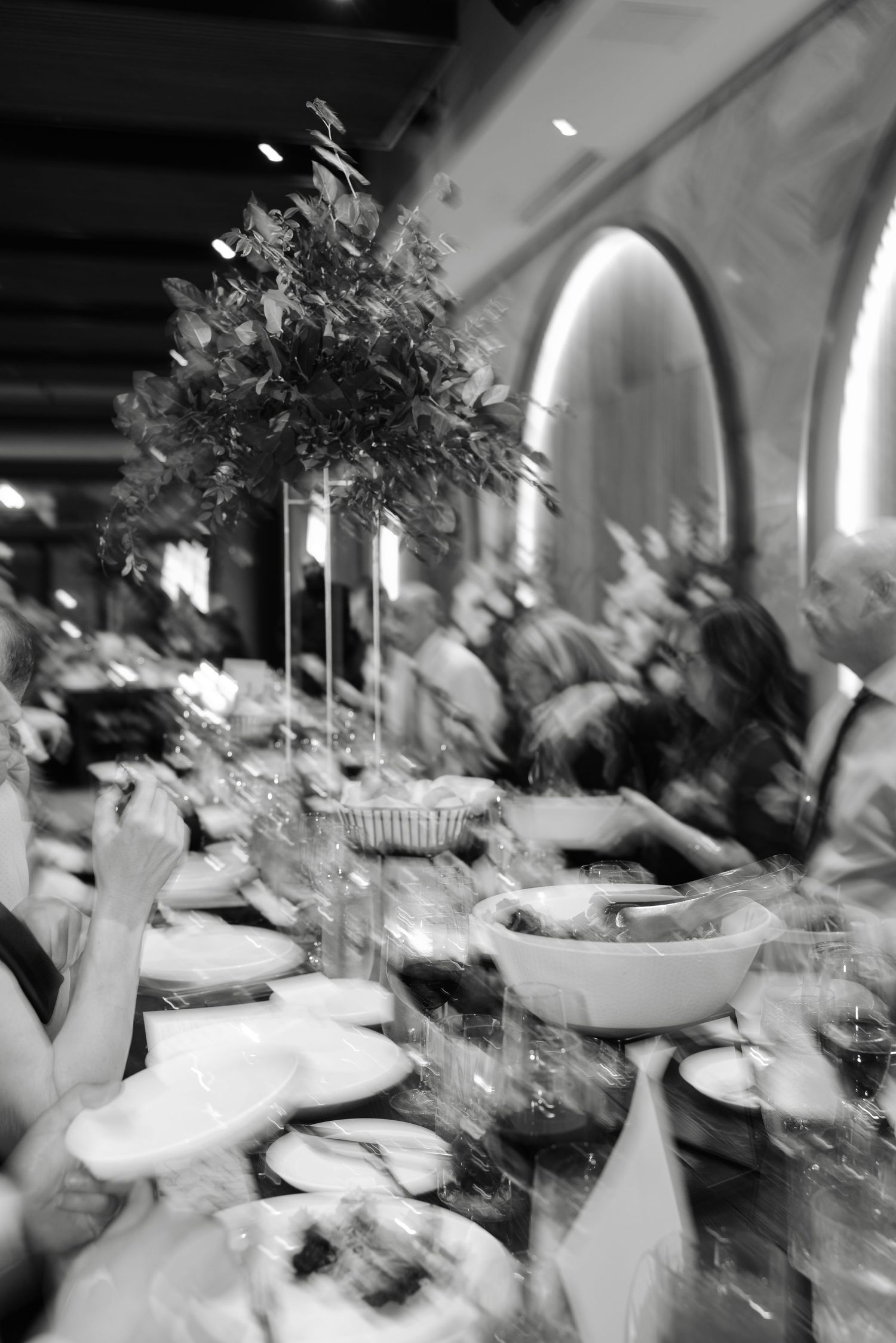 Black and white photo of guests dining at a long banquet table with floral centerpiece in elegant restaurant.