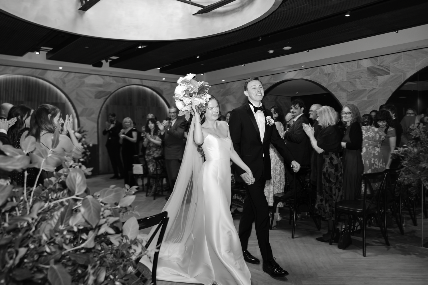 Bride and groom walk triumphantly through cheering wedding guests in elegant black and white reception photo.