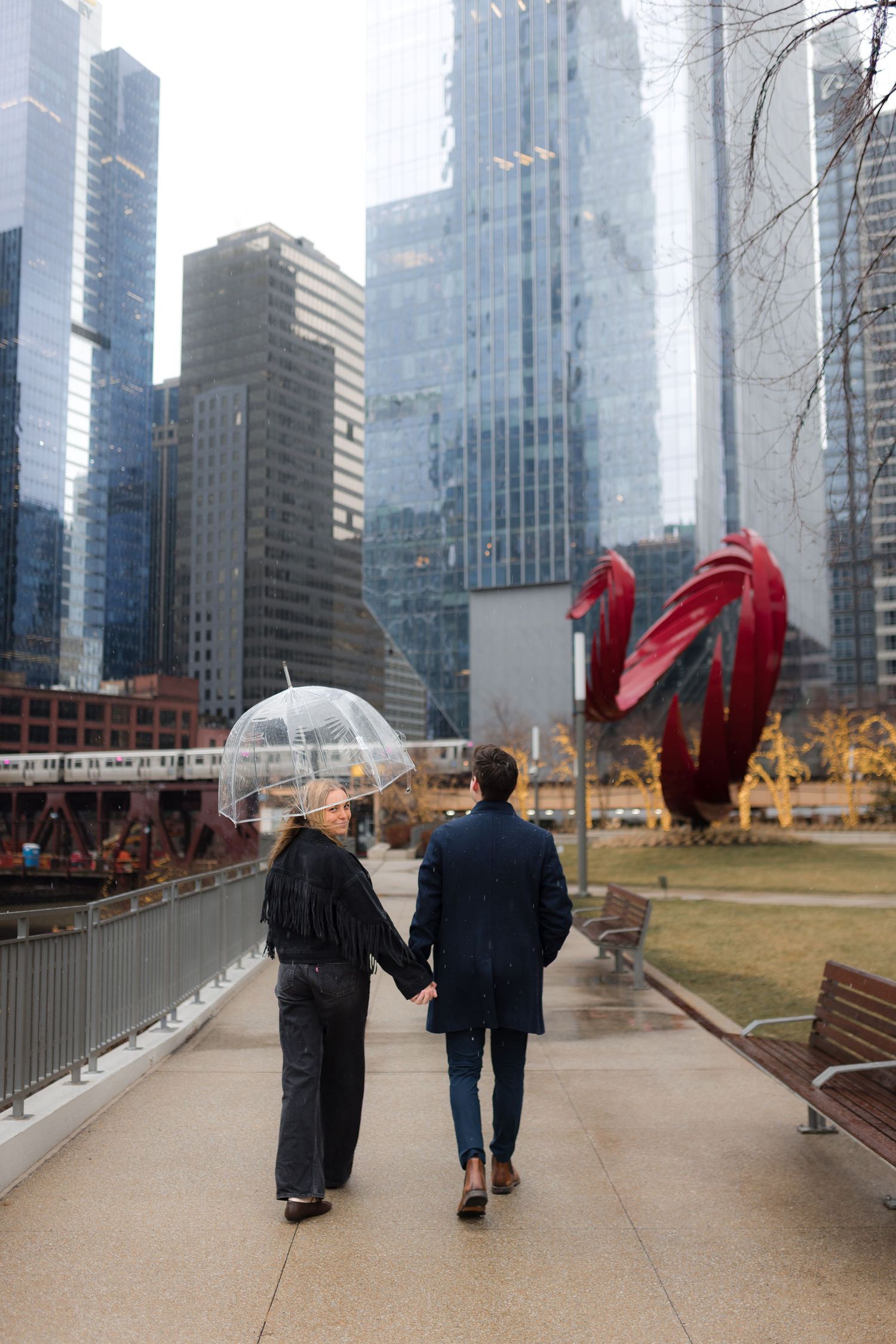 Couple holding hands walking along Chicago Riverwalk with clear umbrella, red sculpture, and city skyscrapers on a rainy day.