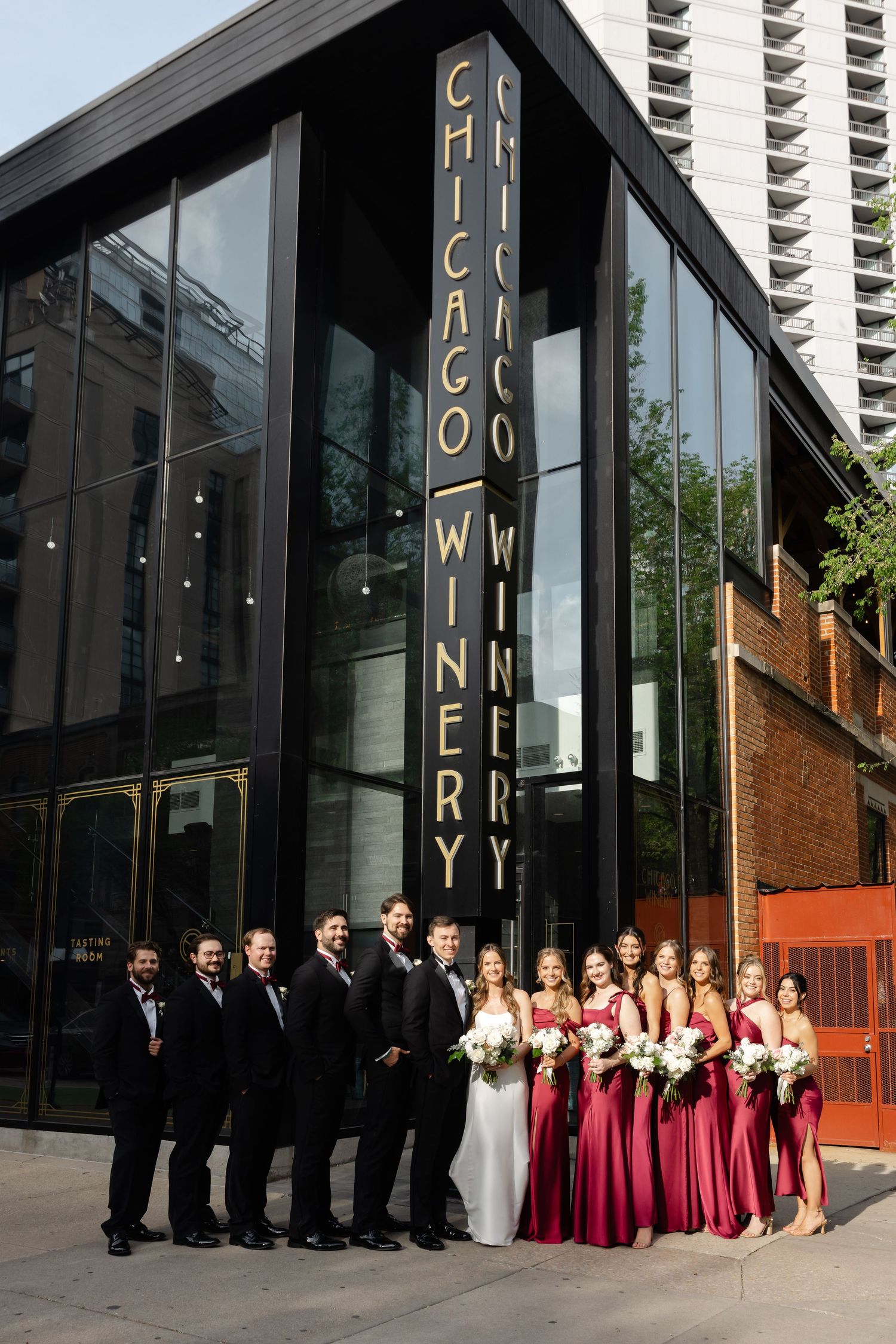 Wedding party posing outside Chicago Winery, groomsmen in black tuxedos and bridesmaids in red gowns.