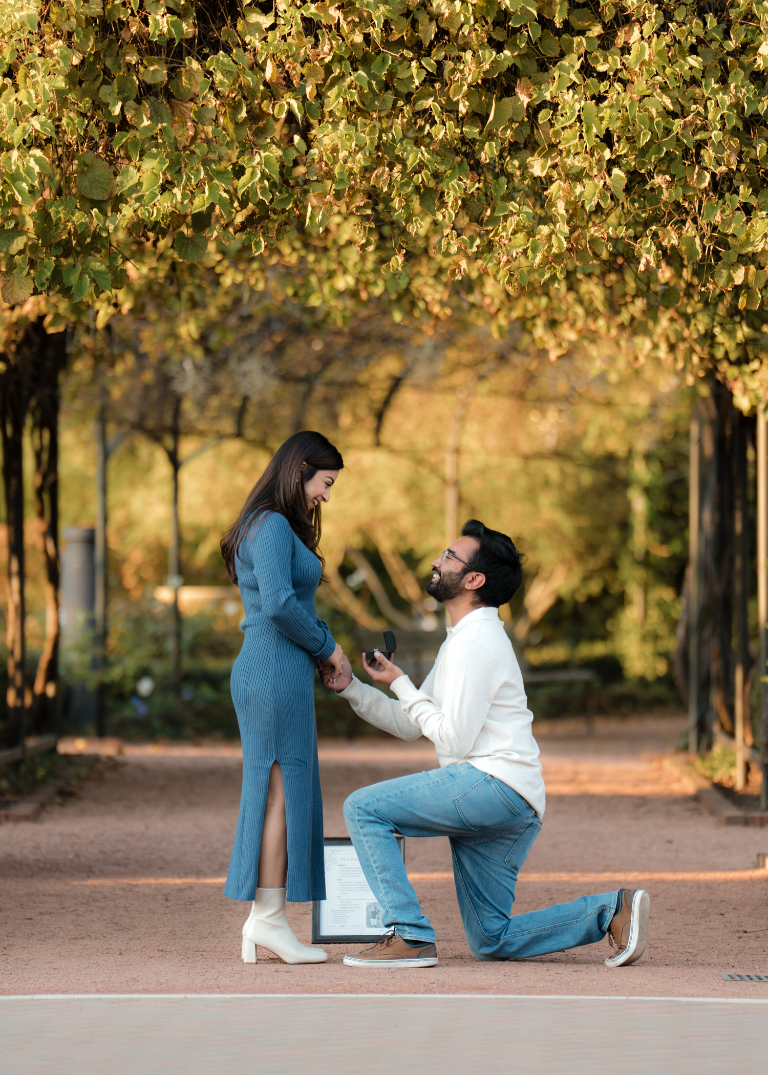 Man proposing on one knee to woman in blue dress under golden autumn trees in a romantic outdoor setting.