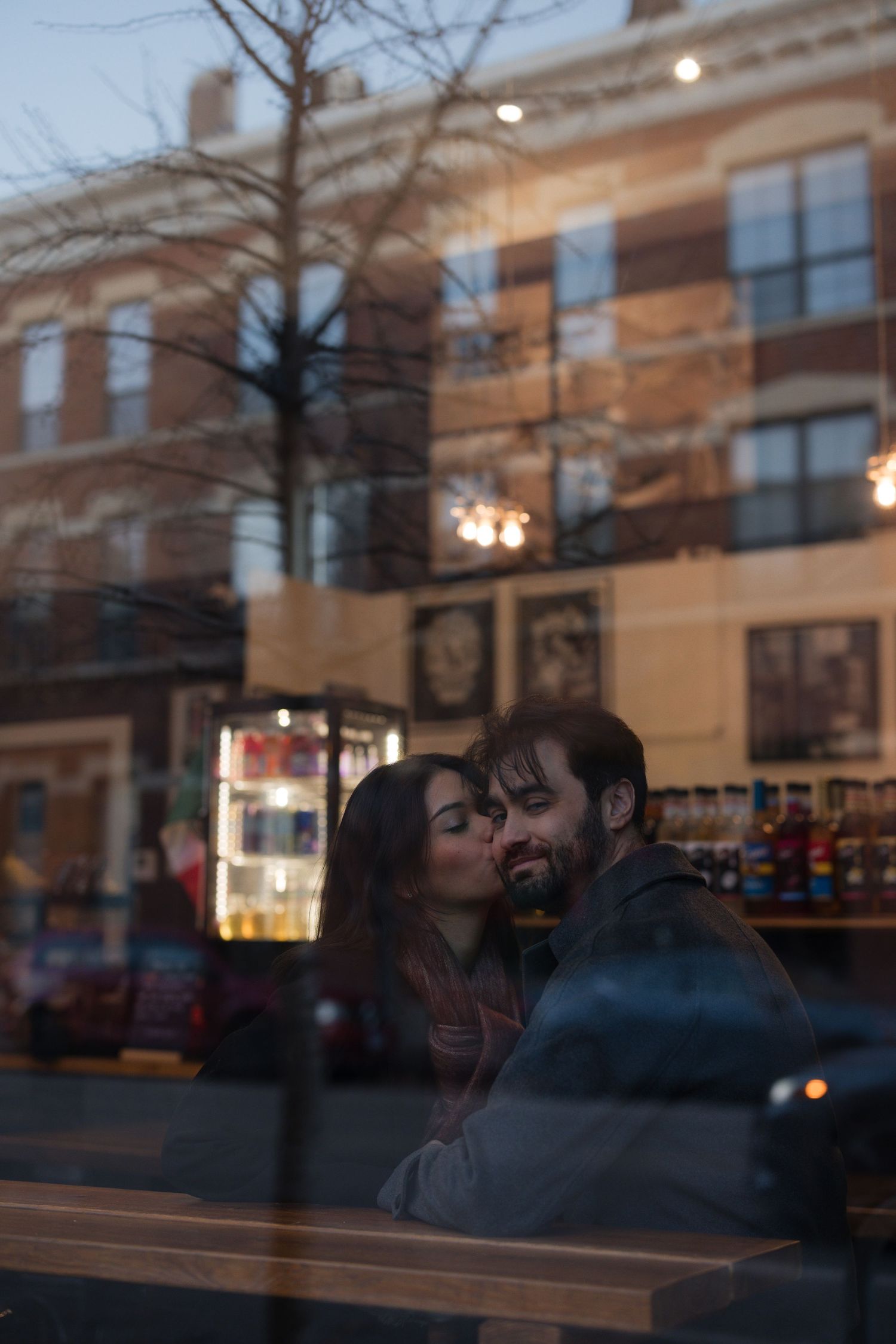 Couple sharing a romantic kiss inside a cafe, captured through a window reflecting a brick building street scene.