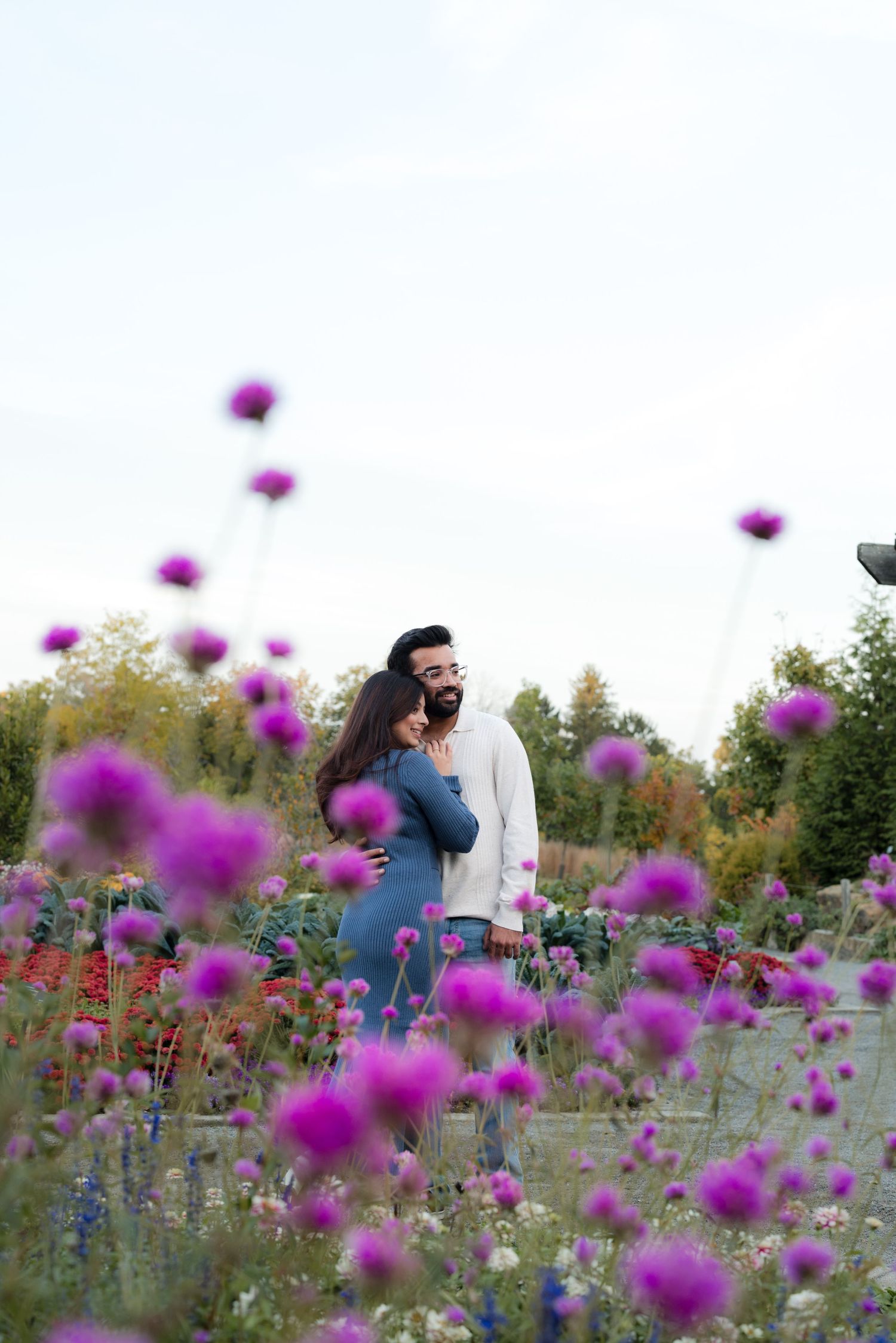 Couple embracing romantically in a vibrant purple wildflower field with lush green trees in the background.