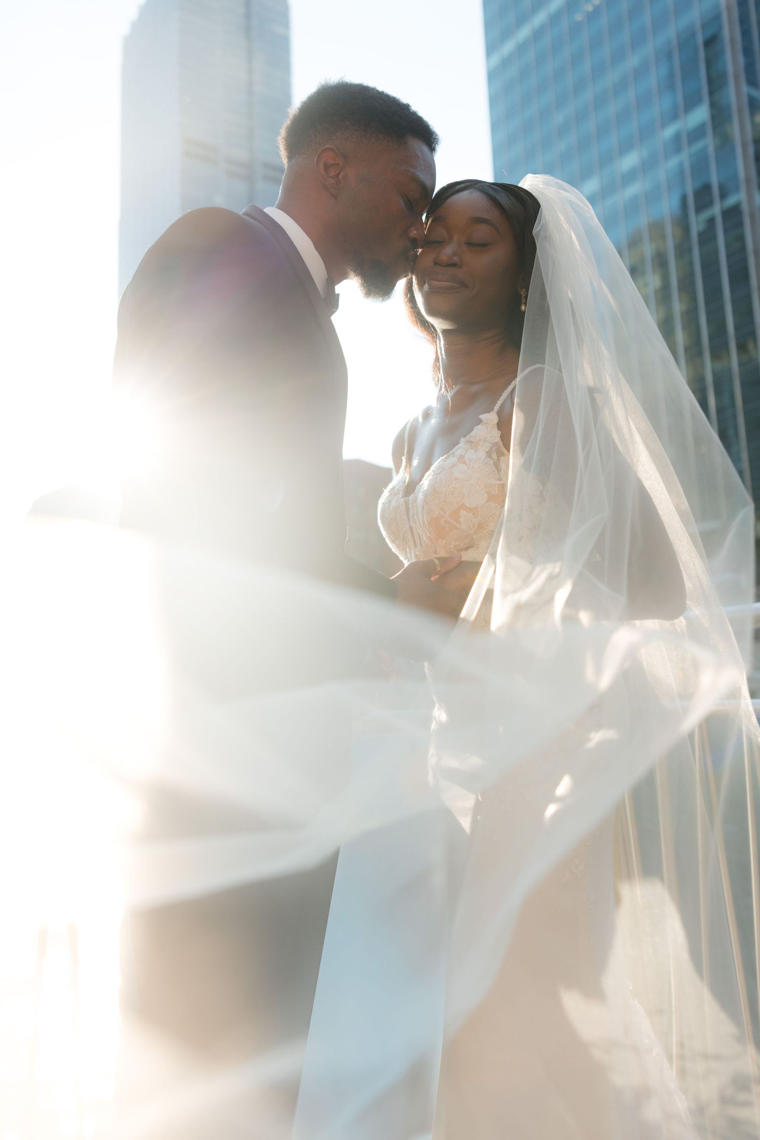 Sun-drenched wedding portraits with flowing veil against modern glass building backdrop.