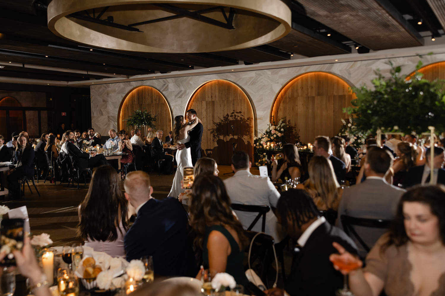 Elegant wedding reception in a grand venue with arched decor, guests seated at tables with gold floral centerpieces.