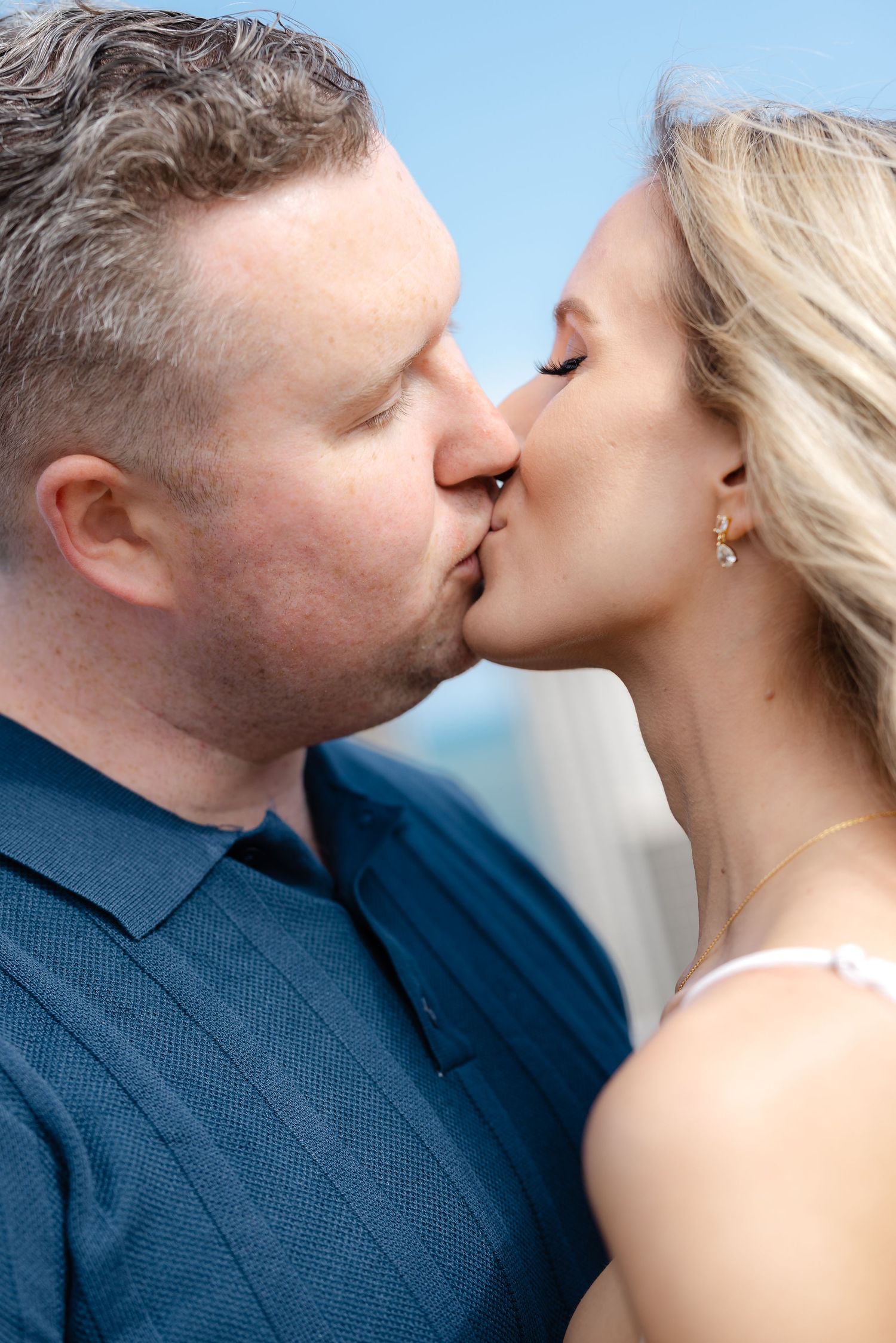 A couple sharing a romantic kiss outdoors, man in navy blue shirt and woman with blonde hair wearing a delicate earring.
