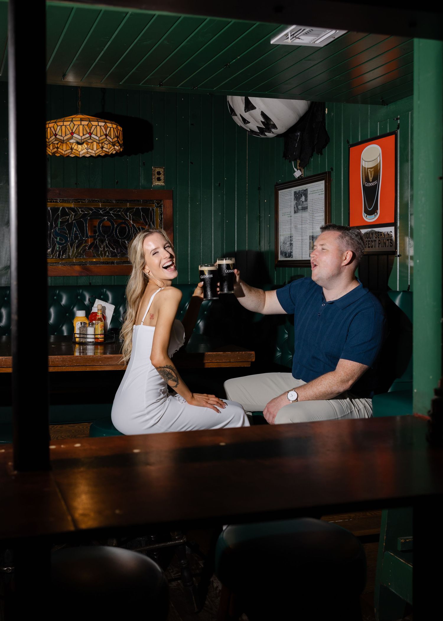 Couple toasting dark beers at a cozy green-walled bar, laughing together in warm ambient lighting.