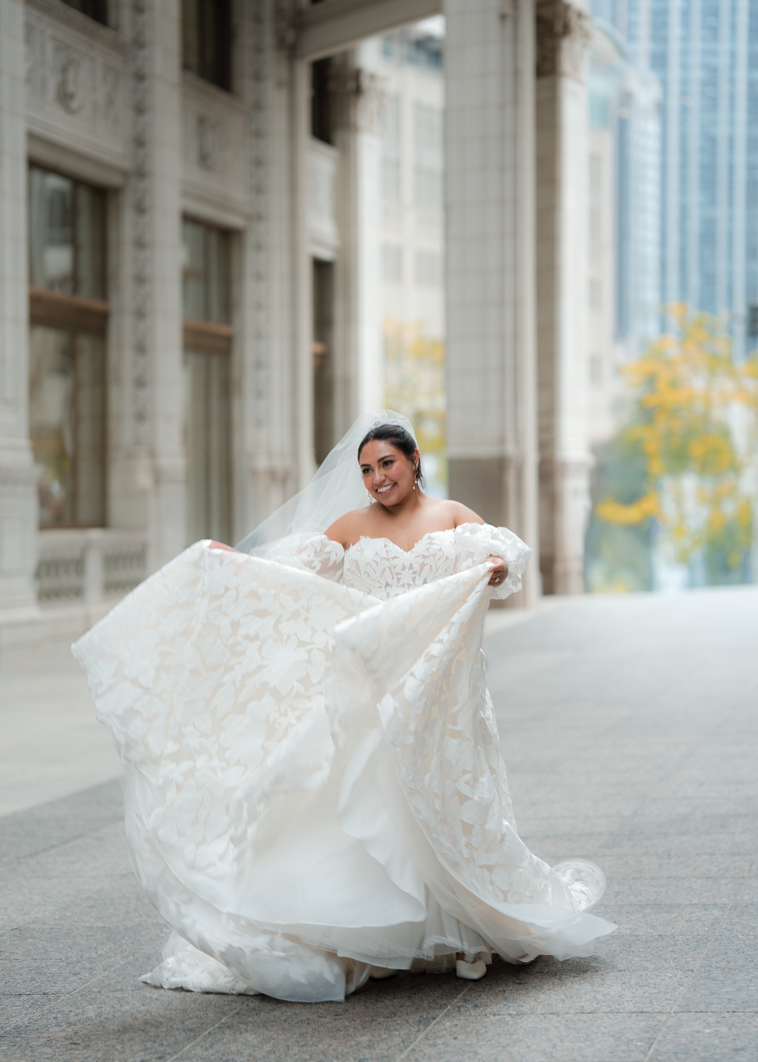 Bride in flowing white wedding gown poses dramatically on urban sidewalk.