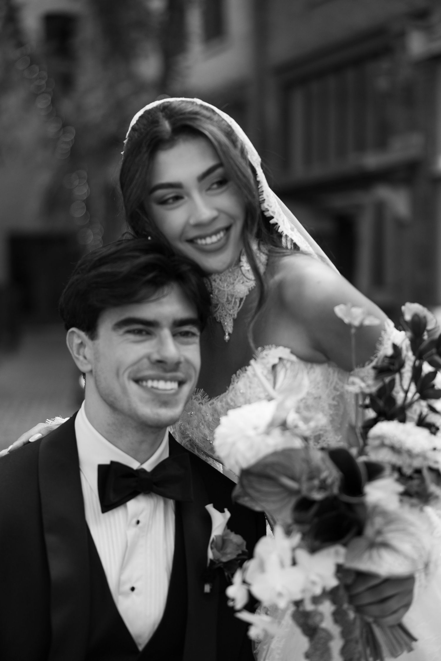 Black and white wedding portrait of smiling bride in veil and groom in tuxedo with bouquet.