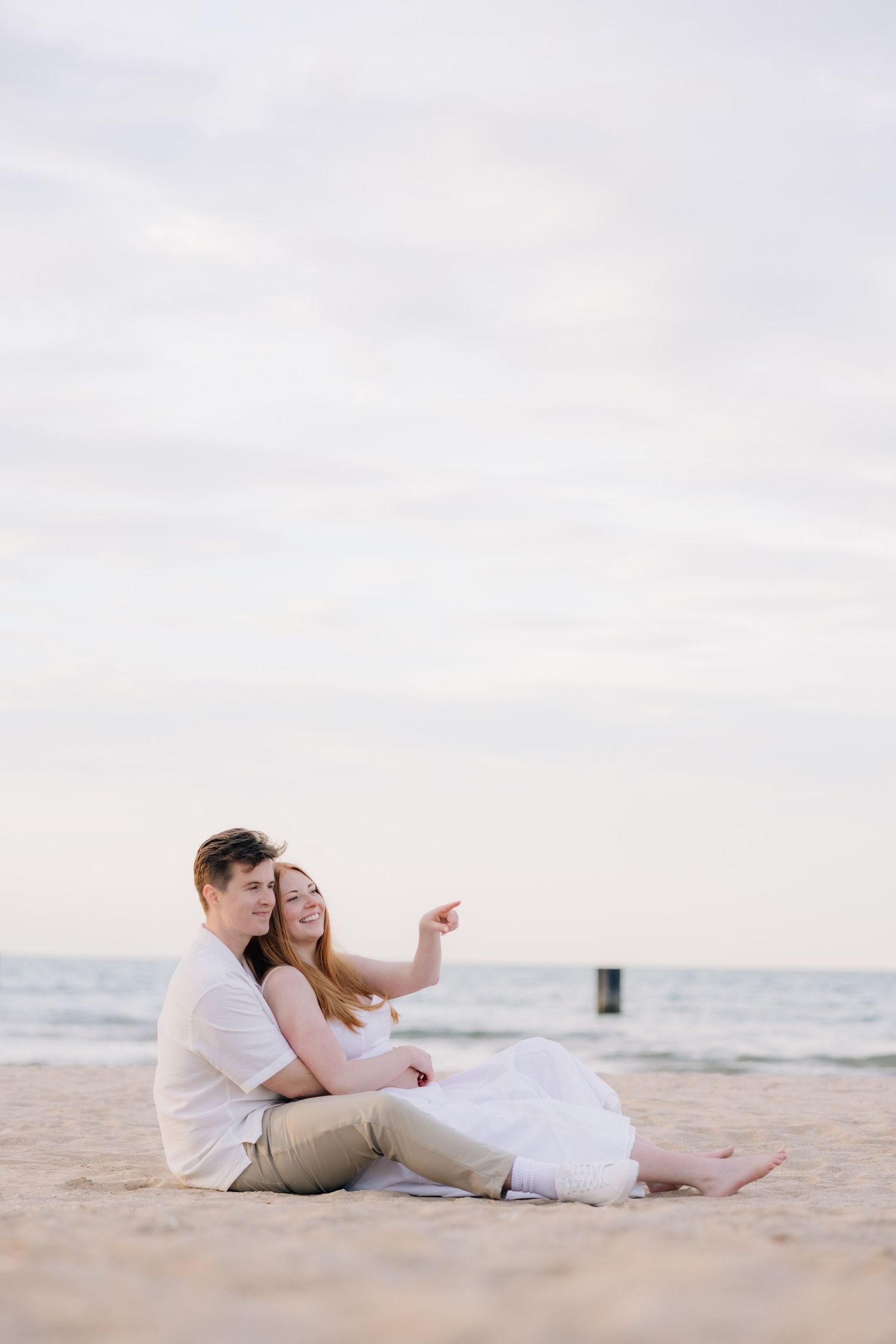 Romantic couple sitting together on sandy beach, woman pointing at horizon, wearing white outfits, overcast sky backdrop.